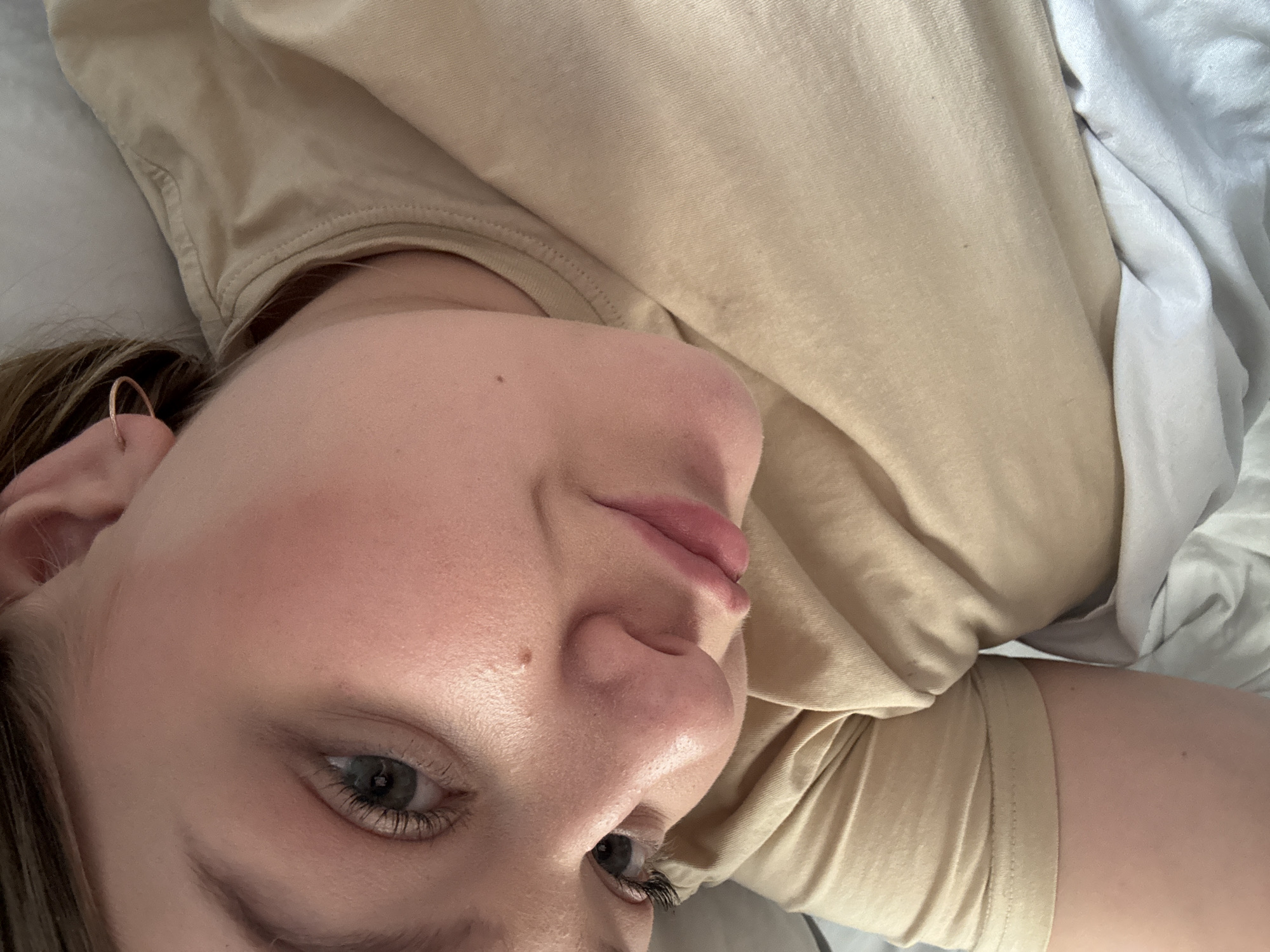 A close-up, high-angle portrait depicts a female individual lying in an indoor setting, presumably a bed, in the city of Chaikovsky, Russia. The subject has fair skin with a subtle pinkish tone on the cheeks and nose, blue-grey eyes with dark eyelashes, and light pink lips that are slightly parted. Brown hair is visible on the upper left side of the frame. A small, thin, copper-colored hoop earring is present in the right ear. A minor mole is located on the upper right cheek. She is dressed in a plain, light beige short-sleeved top. A crumpled white or light grey sheet or blanket is visible beneath her right arm and extending to the lower right corner. The lighting is soft and uniform, illuminating the subject's face.