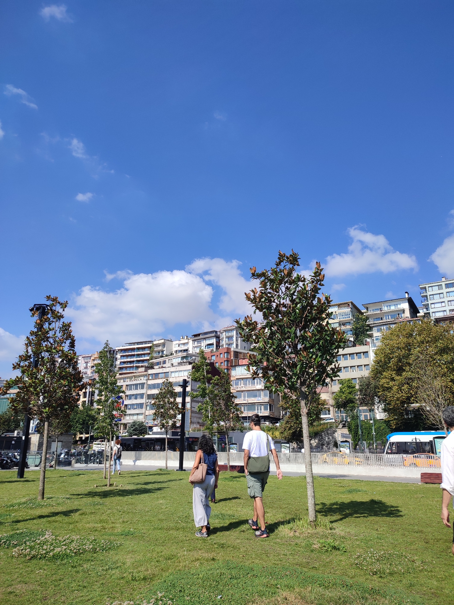 In Beyoğlu, Turkey, a clear blue sky with scattered white clouds provides a backdrop for a cityscape dominated by multi-story buildings constructed on a hillside. In the foreground, a well-maintained grassy park features several young trees. Two individuals are walking away from the camera across the lawn. The person on the left wears a dark top and light-colored pants, carrying a tan bag. The person on the right is dressed in a white t-shirt, green shorts, and athletic shoes, with a green bag slung over their shoulder. Further in the background, a white and blue tram with yellow accents travels along a road bordered by a white fence. The overall scene depicts a pleasant day in a vibrant urban environment.