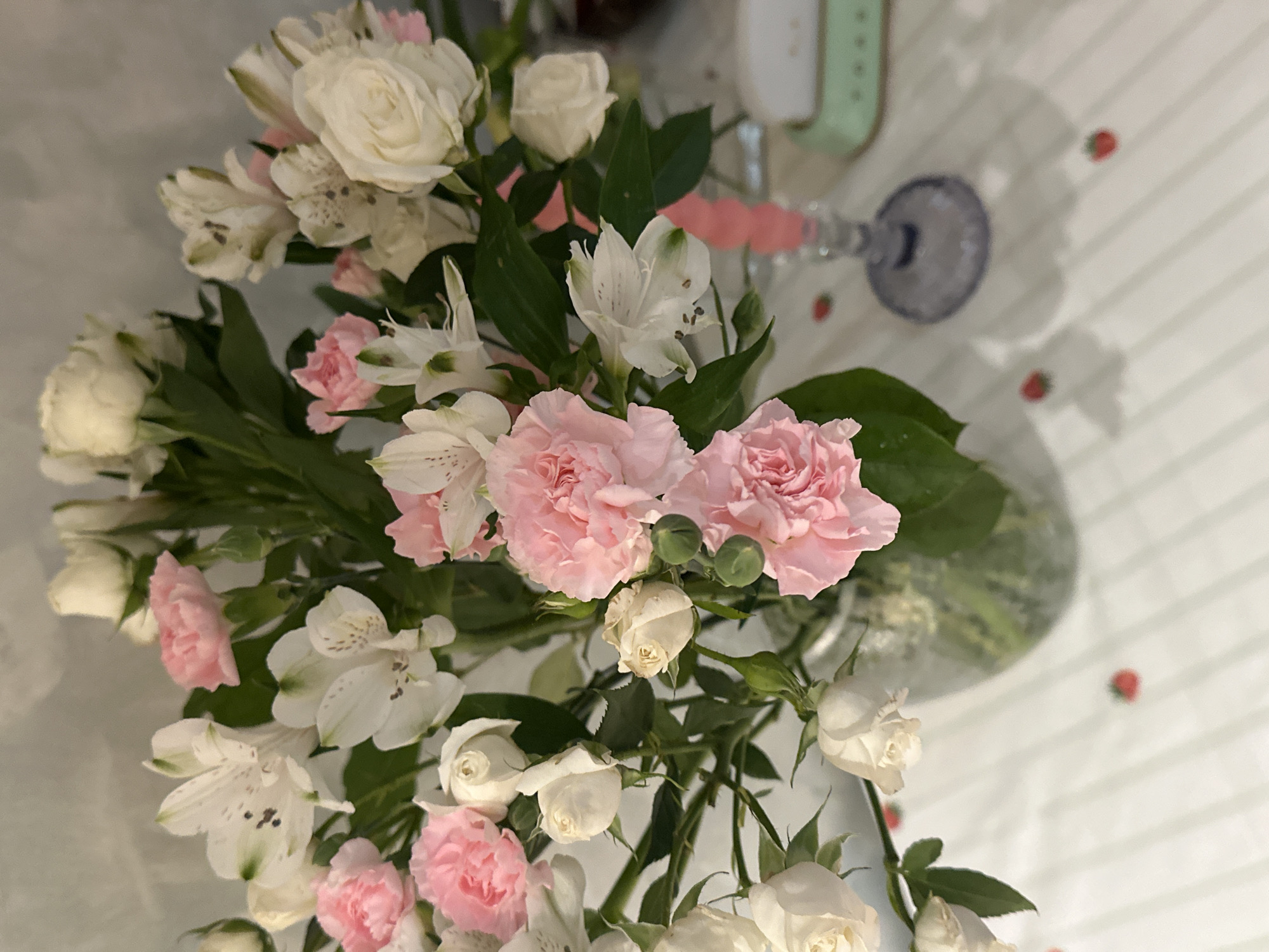 The image shows a bouquet of mixed flowers in a vase on a striped surface. The flowers include pink carnations, white roses, and white alstroemeria. A stemmed glass and a green device (possibly a smartwatch) are also on the surface. Red strawberry-shaped decorations are scattered around. The location is not discernible from the image; therefore, it is not possible to confirm the image was taken in Šušanj, Montenegro.