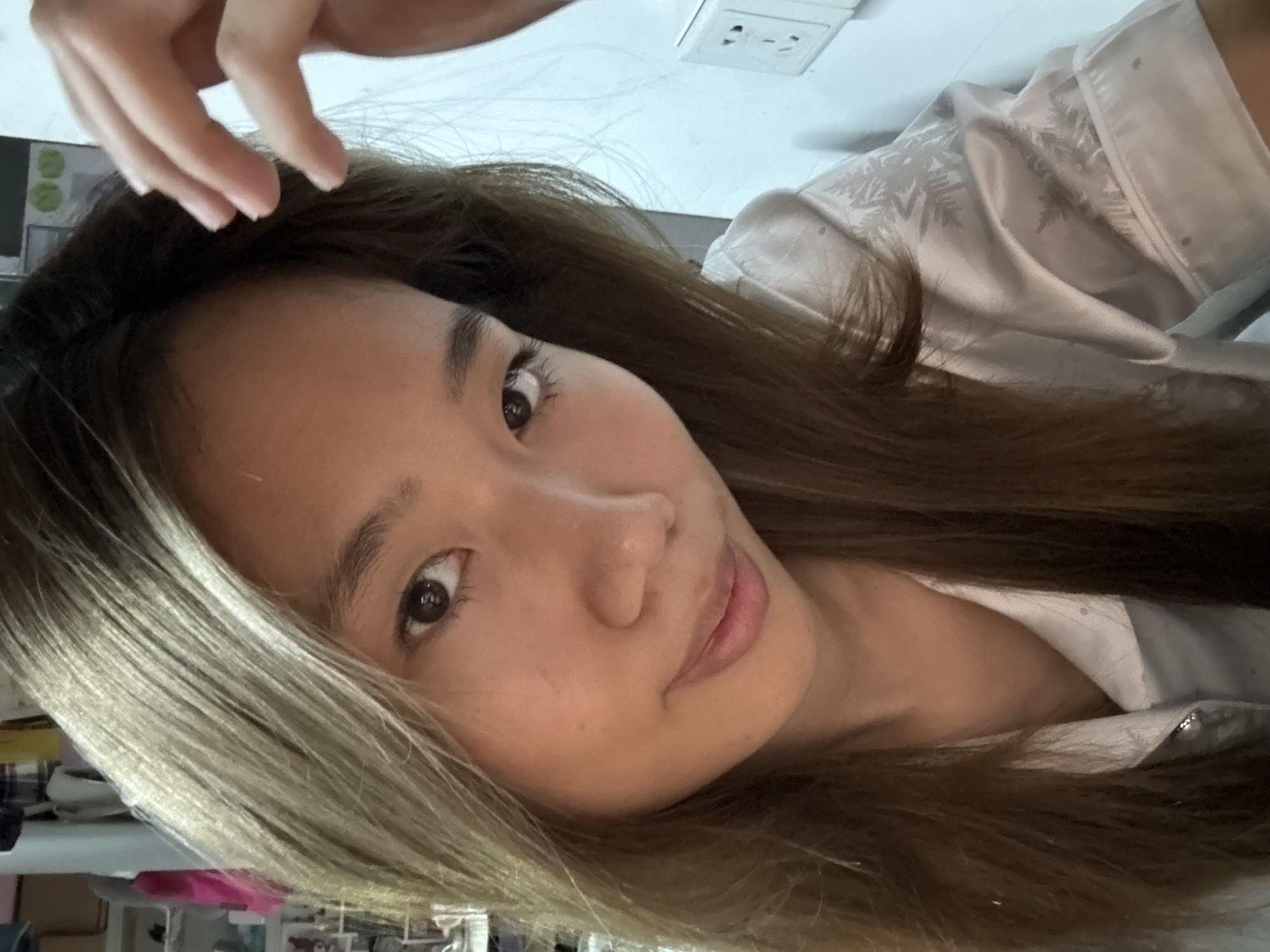 A person with brown and blonde hair is looking at the camera. The person is wearing a white shirt. A hand is partially visible at the top of the frame. A white electrical outlet is in the background. Parts of a desk and chair are in the background. The location is unknown and cannot be determined from the image.