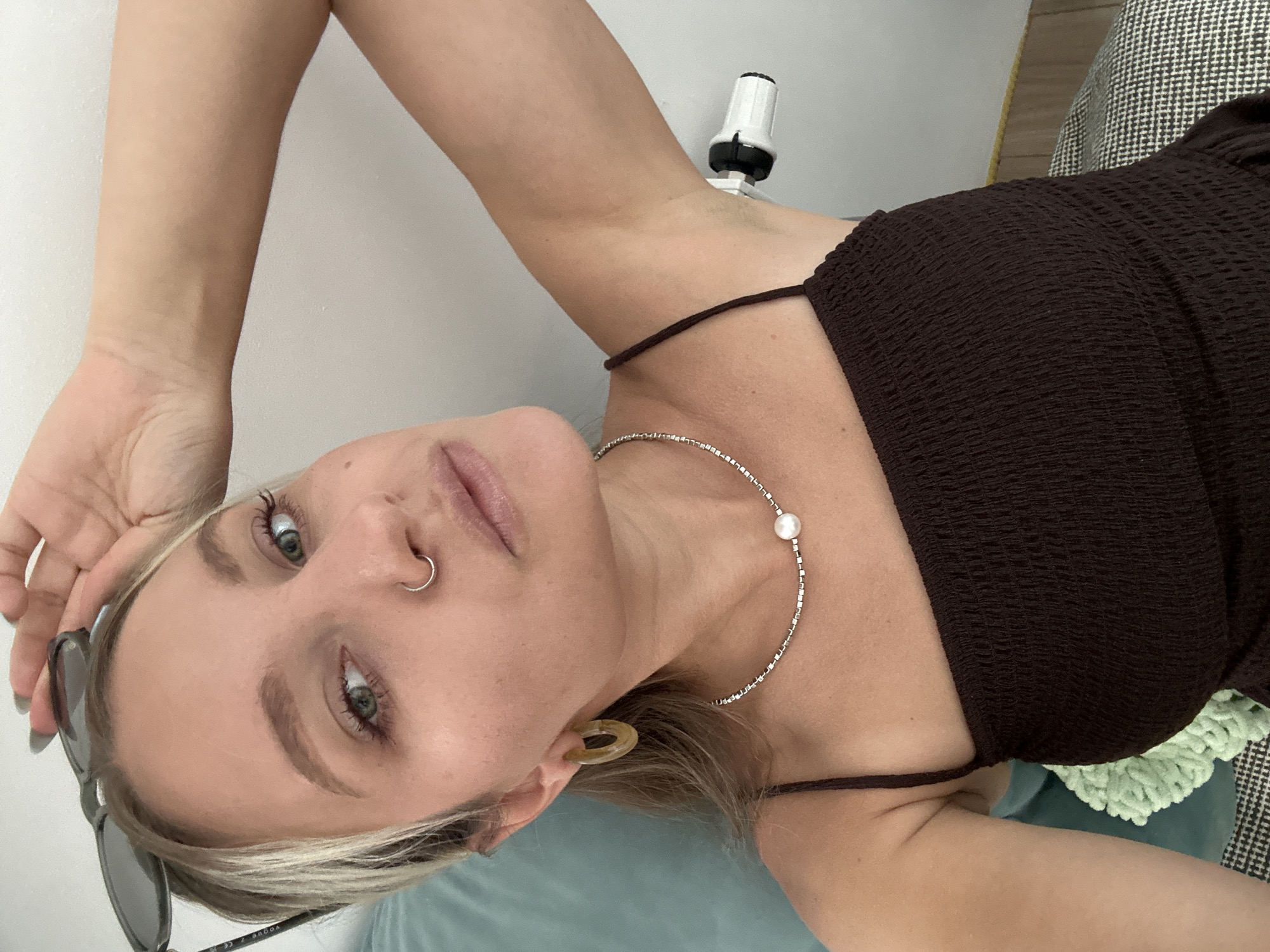 The image shows a medium close-up, high-angle selfie of a young woman with blonde hair, wearing a dark brown smocked top, a pearl necklace, and hoop earrings. She has a nose ring and sunglasses perched on her head. Her right arm is raised, and her hand is touching her head. Her left arm is resting on a teal-colored pillow. A white device with a black cap is attached to the wall behind her left shoulder. The background consists of a white wall and a patterned couch. The scene is indoors. There is no information about the location in Vsevolozhsk, Russia.
