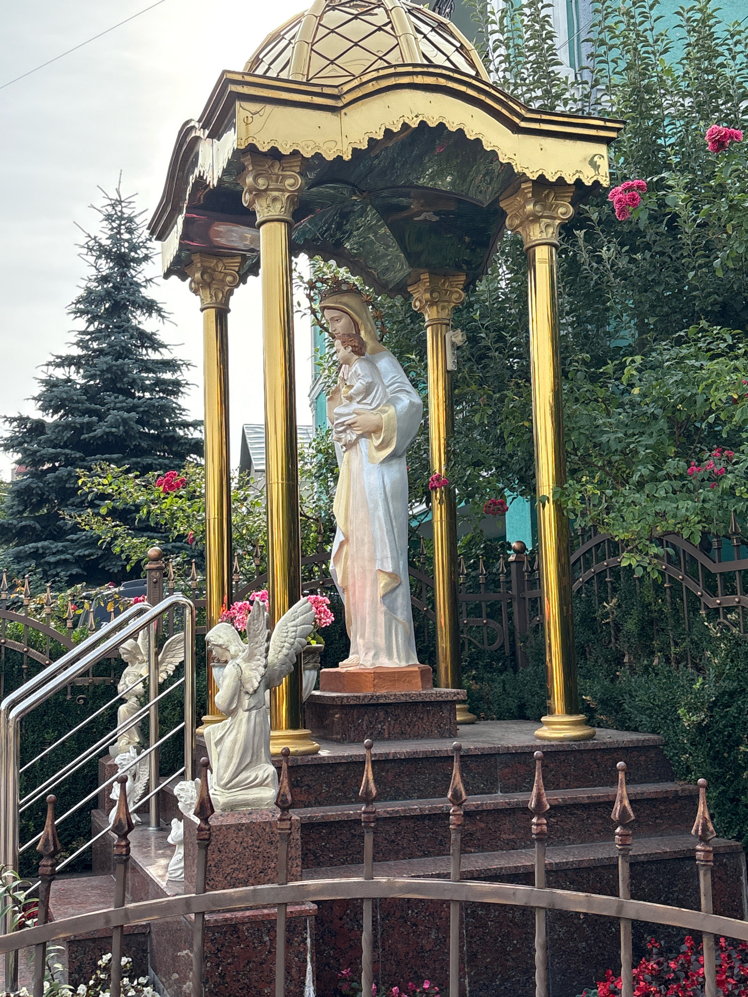 The image shows an outdoor shrine with a statue of the Virgin Mary holding the infant Jesus, standing within a golden gazebo supported by four ornate columns. The gazebo has a domed roof and intricate gold detailing. In front of the main statue, on a stepped granite platform, are several smaller white angel statues, one kneeling and others in standing poses. To the left, a metal railing leads up a granite staircase, with more angel statues visible along the steps. The shrine is surrounded by lush green foliage and pink roses. Behind the shrine, a tall evergreen tree stands out against a cloudy sky. A decorative metal fence encloses the area. The shrine appears to be located in a residential area, possibly in the city of Ternopil, Ukraine, given the architectural style and the presence of religious iconography commonly found in Eastern European countries.