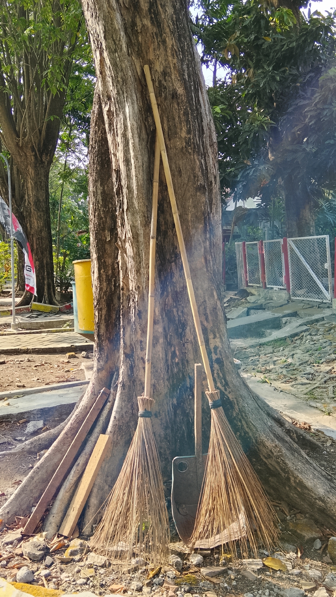 In the city of Tlogobendung, Indonesia, a large tree with prominent roots stands prominently. Leaning against its trunk are two traditional brooms made of dried grass and bamboo handles. Beside the brooms rests a shovel with a broad, flat blade. To the left of the tree, leaning against its roots, are several wooden planks of varying sizes. In the background, a yellow cylindrical trash bin is visible. Further back, a white metal fence with red accents encloses an area. The ground around the tree is covered with a mixture of dirt, small stones, and dried leaves, indicating an outdoor setting. The scene appears to be an unadorned utilitarian space, possibly a maintenance area or a simple outdoor workspace.