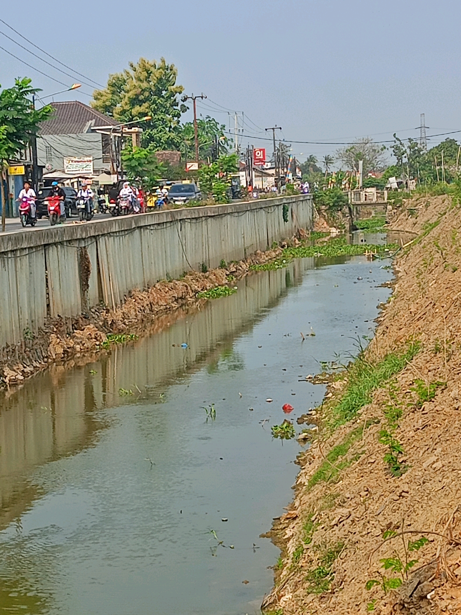 The image depicts a canal running through an urban area in Sriamur, Indonesia. The canal has concrete walls on one side, with a road above where several people are riding motorcycles. The other side of the canal is a steep, unpaved embankment covered in dry soil and sparse vegetation. The water in the canal appears murky and contains some floating debris and aquatic plants. In the background, there are buildings, trees, and utility poles, suggesting a developed area. The sky is clear and bright.