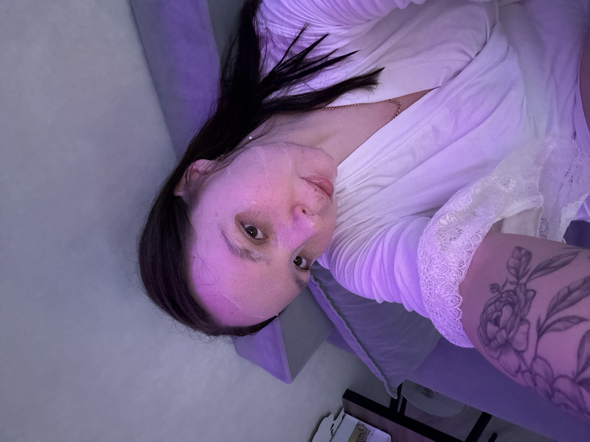 The image shows a female individual in an indoor setting, illuminated by a pervasive purple light source. The specific location within Chaikovsky, Russia, cannot be determined from the visual information provided. The individual, appearing to be in a reclined or semi-reclined position, is looking directly into the camera. Her face has a translucent sheet mask applied, visible as faint lines and a slightly glossy texture on her skin, which also exhibits freckles or minor marks. She possesses long, dark hair, partially visible. She is dressed in a white robe featuring a V-neckline and white lace trim on the sleeve. A gold-colored chain necklace is visible around her neck. A prominent grey-toned floral tattoo, depicting multiple blooming flowers and leaves (resembling peonies or roses), is located on her right forearm/bicep area. The background includes a plain, light-colored wall or headboard, a pillow, and a blurred dark piece of furniture, possibly a table, to the left.