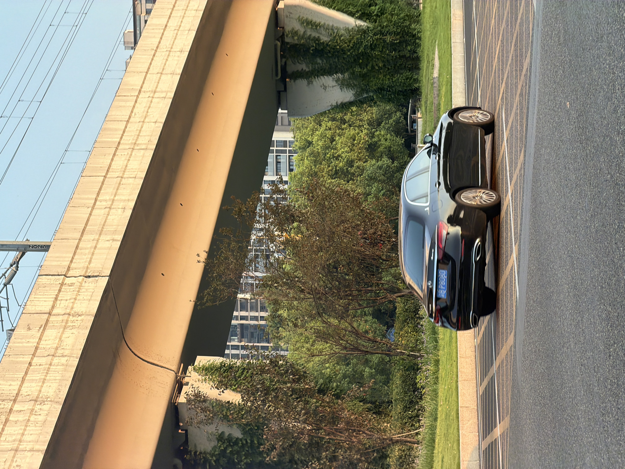 A black Mercedes-Benz sedan is parked on an asphalt road with yellow and white striped markings, indicating a parking area. The car is facing away from the viewer, with its rear towards the camera. In the background, a large concrete overpass casts a shadow over the scene. Green trees and bushes line the area between the road and a row of modern buildings. The sky is clear and light blue. The scene is lit by the warm glow of what appears to be late afternoon or early morning sunlight. The location is identified as Jiangjin, China.