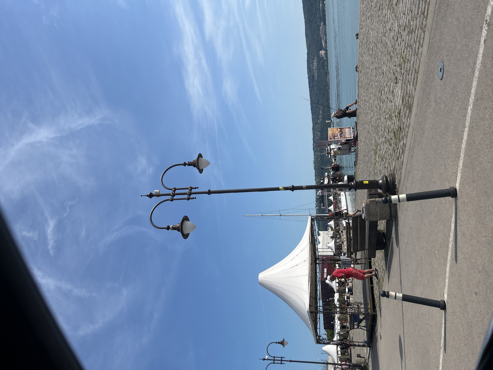 This daytime image captures a scene at the Port of Varna, Bulgaria, specifically along a promenade adjacent to the Black Sea.
The foreground consists of a grey asphalt road with a single white dividing line running horizontally. Two black bollards, each with a white stripe, are positioned on the asphalt, one leaning slightly left and the other slightly right. A circular metal plate is embedded in the asphalt near the bottom right corner.
Beyond the asphalt is a paved area featuring cobblestones or similar patterned pavers. A prominent, large white conical tent structure with an open metal frame stands on the left side of this paved area, under which benches are visible. Further down the promenade, several smaller white umbrella-like structures or tents are scattered. To the left of the large tent, another streetlight is partially visible. A man in a red shirt and red shorts stands under the large tent, facing right.
A tall, dark metal streetlight with two antique-style lamps on curved arms is centrally positioned, rising from the paved area. A grey concrete bench is situated at its base. Behind the tents, a building with "DOLCE AMARO" partially visible on its facade is present, and a white sailboat mast extends into the sky.
To the right of the central streetlight, two individuals are engaged in fishing along the stone edge of the pier. One person is seated on a dark box or cooler, holding a fishing rod over the water. Another person is seated further left, near a vertical poster board displaying text and images. Boats are docked along the waterfront to the left.
The background features a large expanse of calm, blue water. Beyond the water, a distant, heavily forested hillside with scattered buildings on its slopes is visible under a bright, clear blue sky containing sparse, wispy cirrus clouds. The top left corner of the image is obscured by a dark, angled structure.