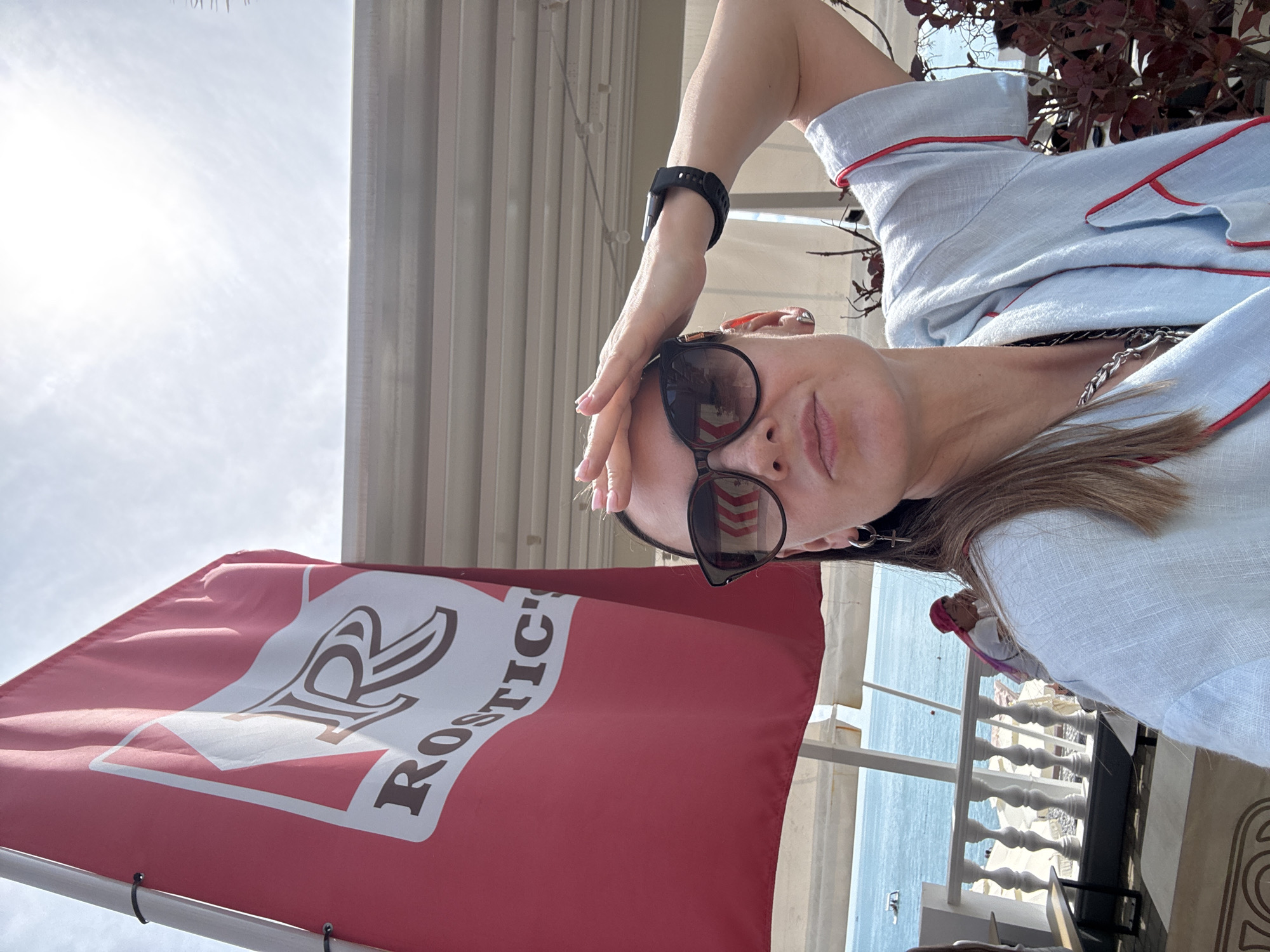 A woman with long brown hair and sunglasses holds her hand to her forehead, shielding her eyes from the sun. She is wearing a light blue shirt with red trim and a silver necklace. In the background, a red flag with a white logo that reads "ROSTIC'S" is visible. Beyond the flag, a beach with white umbrellas and lounge chairs can be seen, along with the sea. A person wearing a pink headscarf is seated at a table near the beach. The scene appears to be set in a cafe or restaurant by the sea, likely in Sochi, Russia, given the branding.