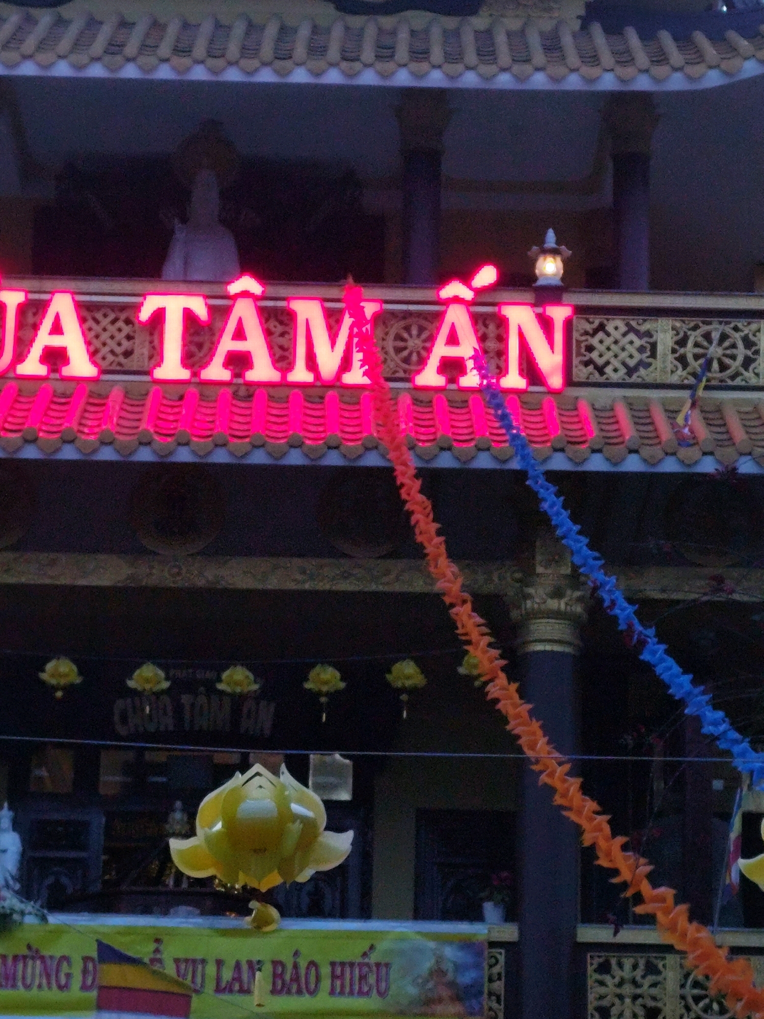 The image shows the facade of a Buddhist temple, likely Chua Tam An, in Da Lat, Vietnam, illuminated by neon pink lettering that reads "CHUA TAM AN" across the top. Decorative lanterns are visible on the roofline, and below the neon sign, a traditional temple railing with intricate patterns is present. Two strings of paper decorations, one orange and one blue, hang down from the upper levels. In the foreground, a large, yellow, lotus-shaped decoration is suspended, and a green banner with Vietnamese text and a flag is partially visible at the bottom. The overall impression is one of a brightly lit, festive temple exterior at dusk or night.