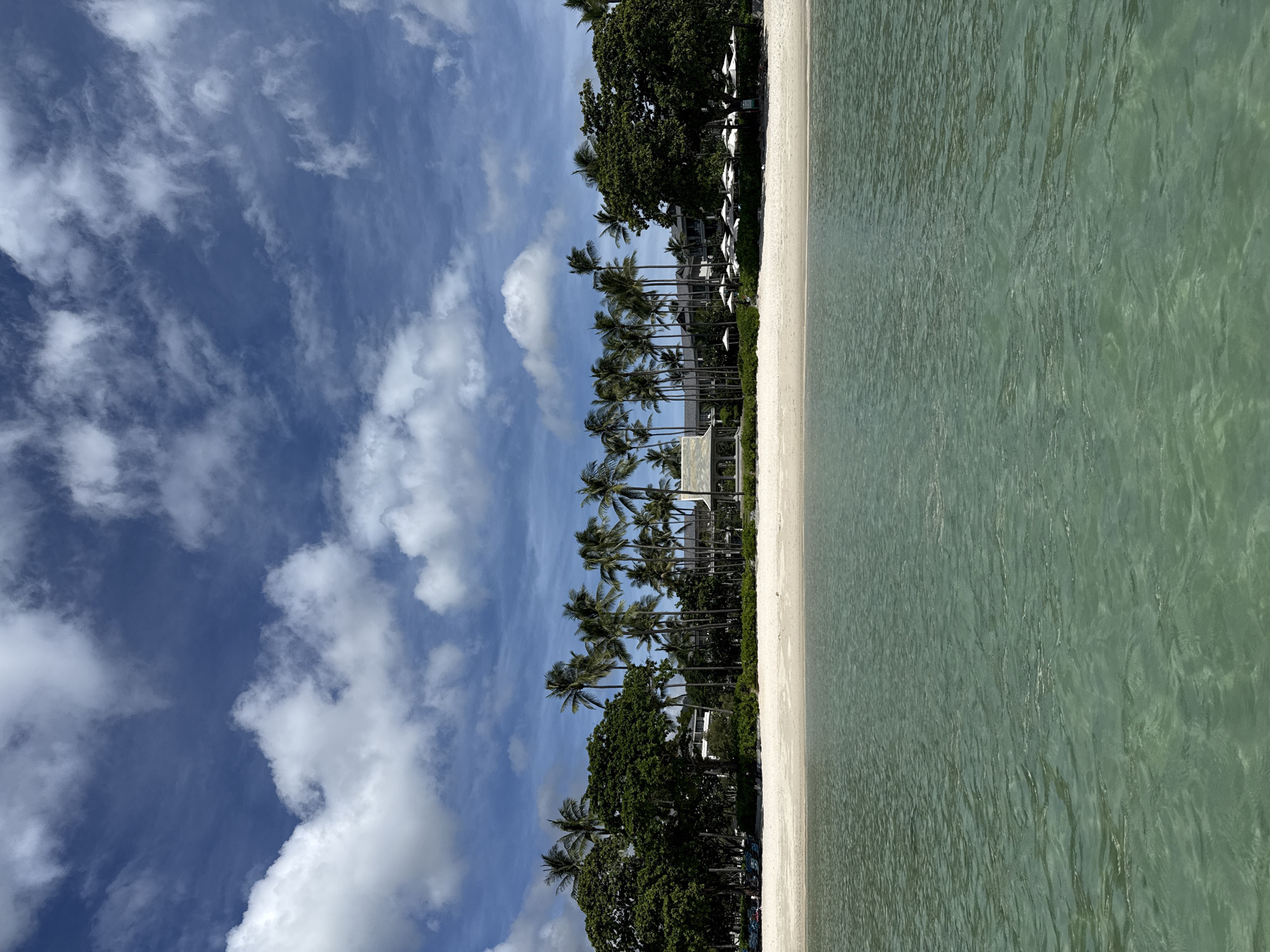 The image shows a tropical beach scene in Baan Chaweng, Thailand. The foreground is dominated by clear, turquoise water with gentle ripples. A sandy beach stretches across the middle ground, lined with lush green trees and tall palm trees. In the background, several buildings, likely part of a resort, are visible behind the vegetation. White umbrellas are set up on the beach, suggesting a relaxation area. The sky above is a vibrant blue with scattered white, fluffy clouds. No people are clearly visible in the image, but beach chairs are present on the sand. The scene conveys a sense of tranquility and a serene tropical atmosphere.