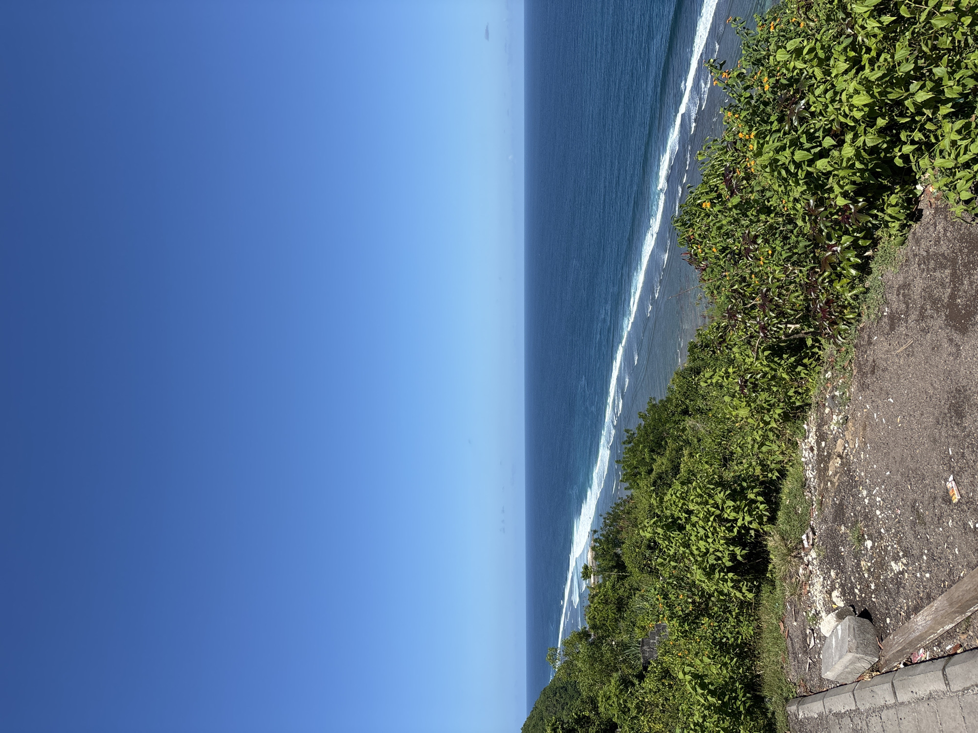 The image shows a coastline view in Kuta Selatan, Indonesia. The sky is clear and blue, occupying the left half of the frame. A body of water, likely the ocean, fills the space between the sky and the right edge of the frame. White waves can be seen breaking along the shoreline. Lush green vegetation grows along what appears to be a cliff or elevated area on the right side of the frame. A paved surface, possibly a road or pathway, runs along the edge of the vegetation.