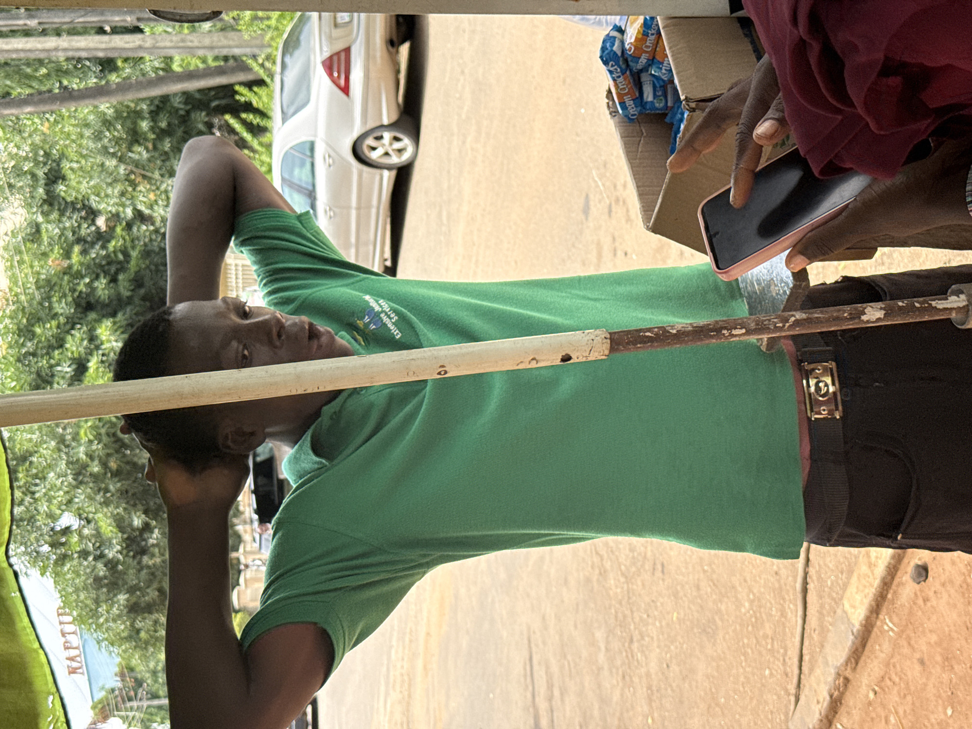 An African man wearing a green polo shirt with a logo and black pants with a belt is standing outdoors, possibly in Abuja, Nigeria. His hands are behind his head, and he appears to be speaking or reacting to something. A white pole is in front of him. To the right, a box of crackers is visible, and a hand is holding a pink phone, suggesting an interaction or transaction. In the background, trees and a light-colored sedan are visible, indicating an urban or semi-urban environment. The ground appears to be a dirt road.