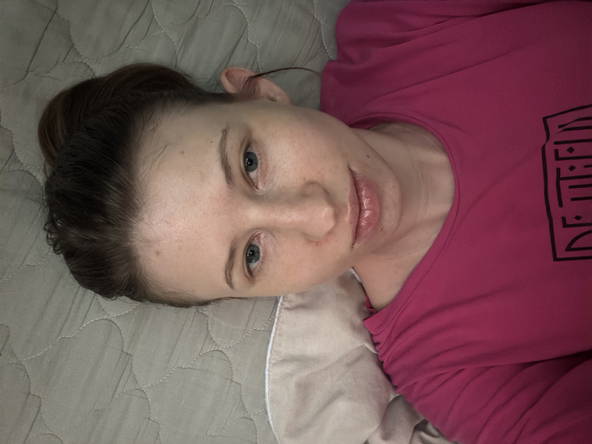 A young Caucasian woman with light skin and brown hair pulled back in a bun is lying on a textured gray surface, possibly a mattress, indoors. She is wearing a bright pink t-shirt with a black graphic design on the chest. Her eyes are open and directed towards the camera, and her lips are slightly parted. A beige pillow is visible to her left. The scene appears to be a casual, personal setting. The image does not provide information to determine the specific location within Dongducheon-si, South Korea.