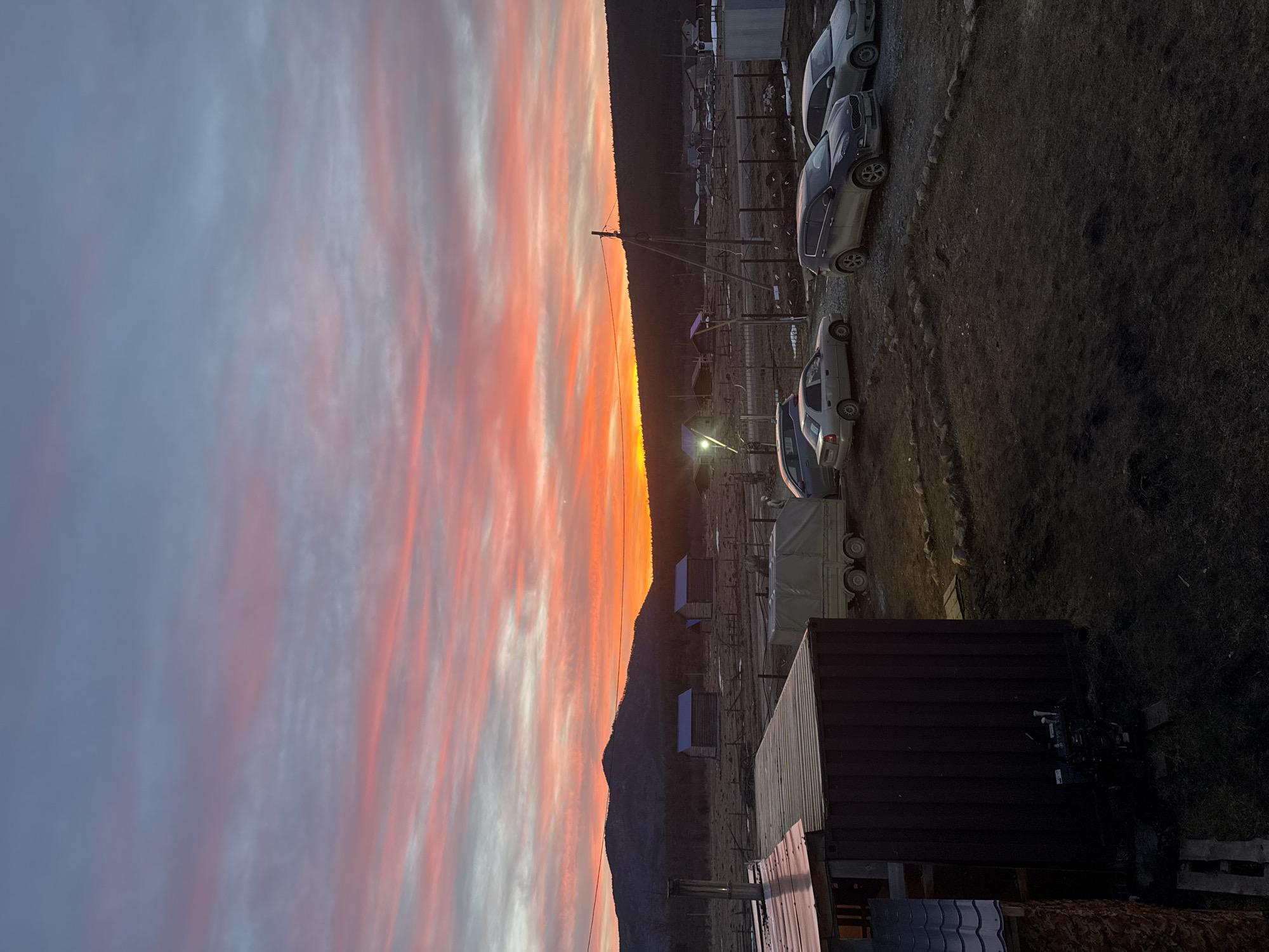 The image depicts an outdoor scene in Tyulyuk, Russia, at either sunrise or sunset. The sky is dominated by vibrant orange and red hues along the horizon, transitioning upwards through pink to pale blue-grey, with visible horizontal cloud streaks. A dark, forested mountain range is silhouetted across the background.
In the left foreground, the dark, corrugated metal side and partial roof of a building or large container are visible. A small, dark-colored engine or generator is situated on the ground near its base. The immediate ground consists of bare, uneven dark earth with sparse vegetation and scattered rocks.
The midground features a dirt and gravel area. A light-colored, two-wheeled utility trailer covered with a tarp is parked on the left. To its right, three vehicles are present: a light-colored sedan, a dark-colored SUV (likely a Kia Sportage), and another light-colored sedan. Further back, a series of fences, constructed from both wood and wire, delineate sections of the landscape. Two rustic wooden cabins with prominent bright blue roofs stand in a clear field. Multiple utility poles with overhead power lines are visible, with one prominent pole in the mid-right portion of the frame. In the distance, a cluster of small structures, indicating a village or settlement, is visible, with one building displaying a distinct light source. No human figures are visible in the scene.