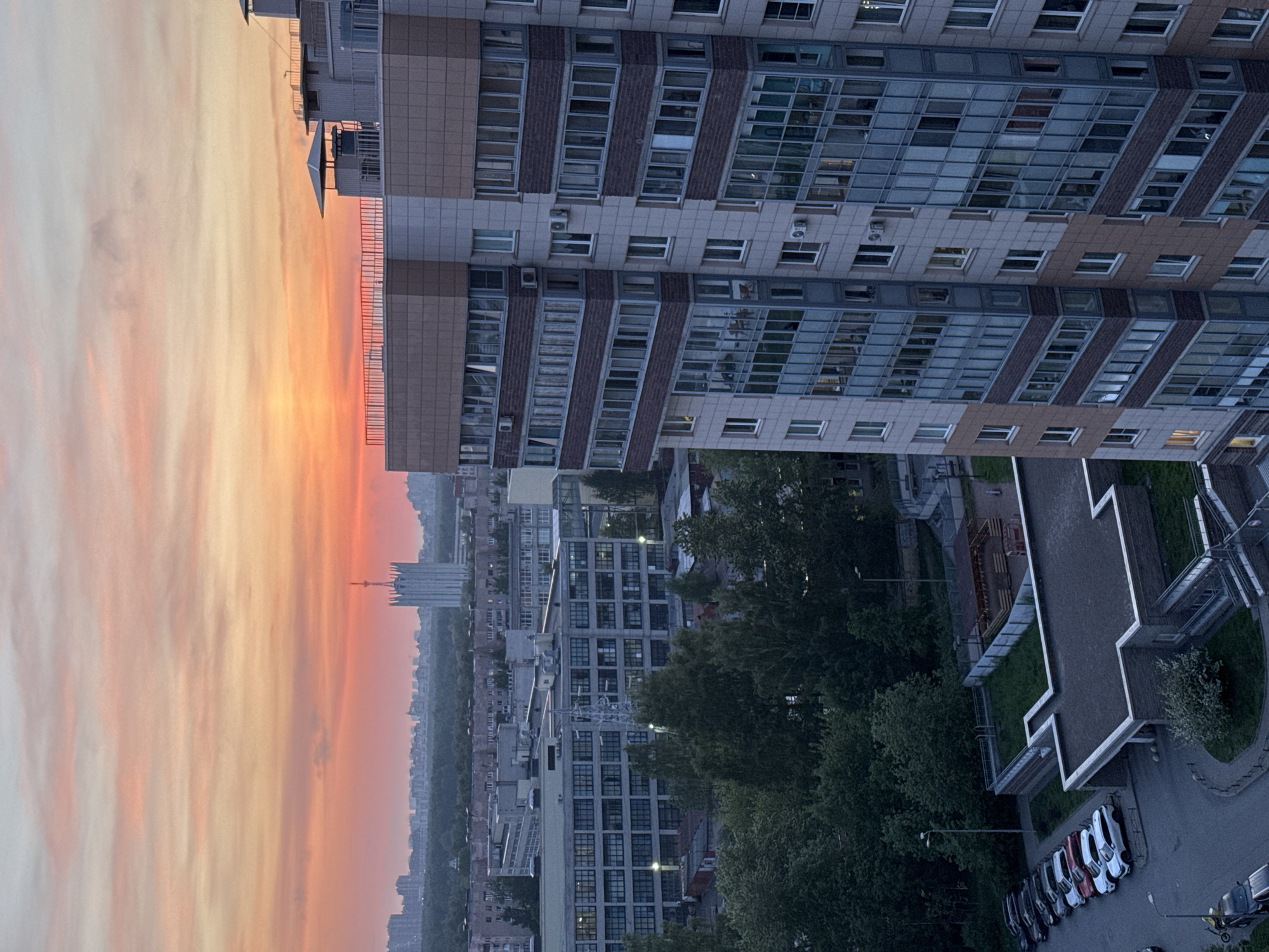 The image depicts a view over Ladozhskoe Ozero, Russia, during a sunset. The sky is colored with orange and yellow hues. In the foreground, a tall modern building with multiple windows and gray and brown paneling dominates the right side. Below the building, there's a courtyard with trees, a walkway, and a row of parked cars. In the distance, more buildings and a tower can be seen on the horizon. The overall environment suggests an urban setting during twilight.