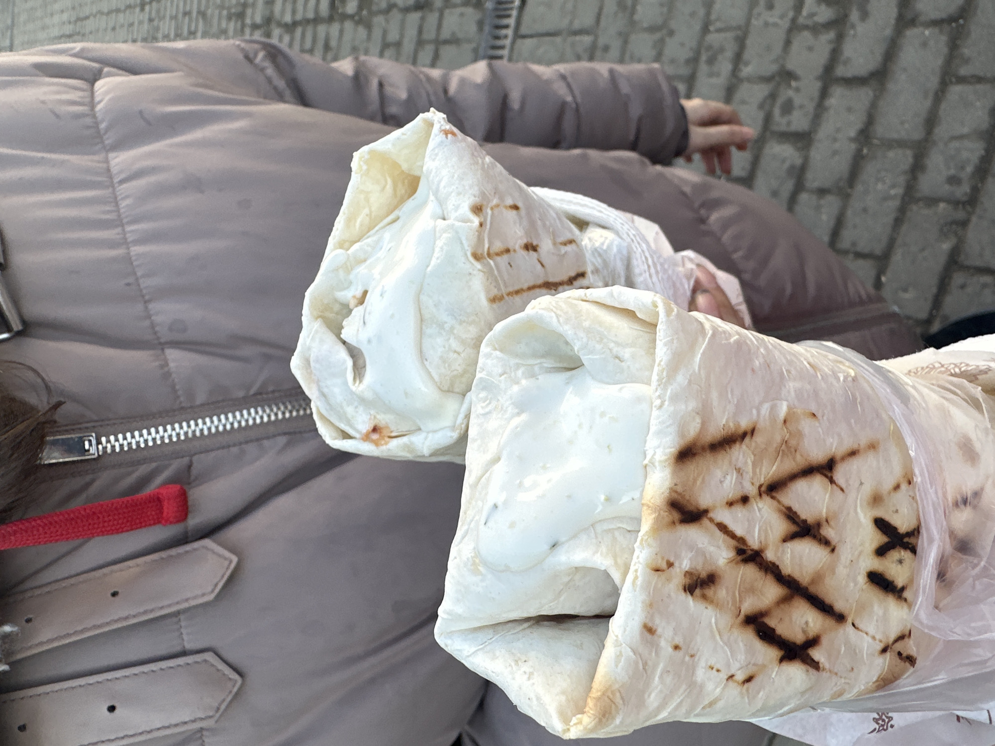 Here is a description of the image:
Two shawarmas are held over a puffy brown jacket. The shawarma on the right has been grilled and is partially wrapped in plastic. The shawarma on the left is not grilled. The scene appears to be outdoors with a paved walkway or street visible in the background. The zipper and belt details of the jacket are visible. The location is Moscow, Russia.