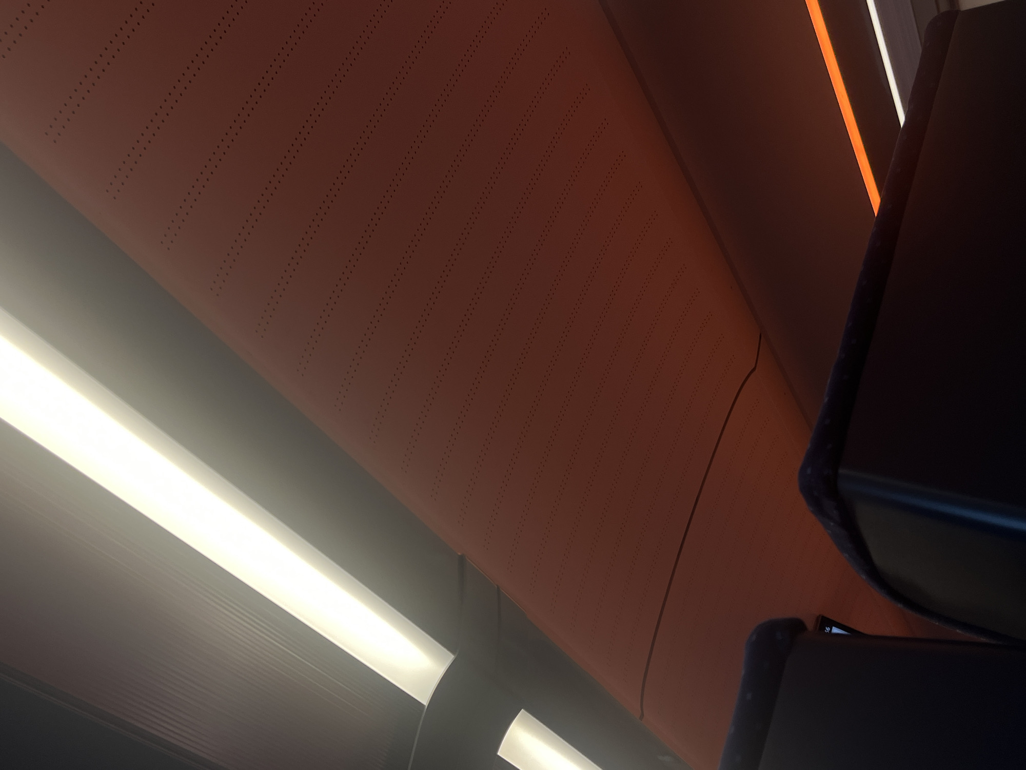 The image depicts the interior of a vehicle, likely a train or bus, looking upwards. The ceiling features two illuminated strips, one bright white and the other a warmer orange-toned light. A textured, reddish-orange panel with rows of small perforations is prominent, angled across the upper portion of the frame. In the foreground, the dark, upholstered headrests of two seats are visible. The scene is devoid of any people or overt actions. The precise location in Ilmenau, Germany, is not discernible from the visual information provided.