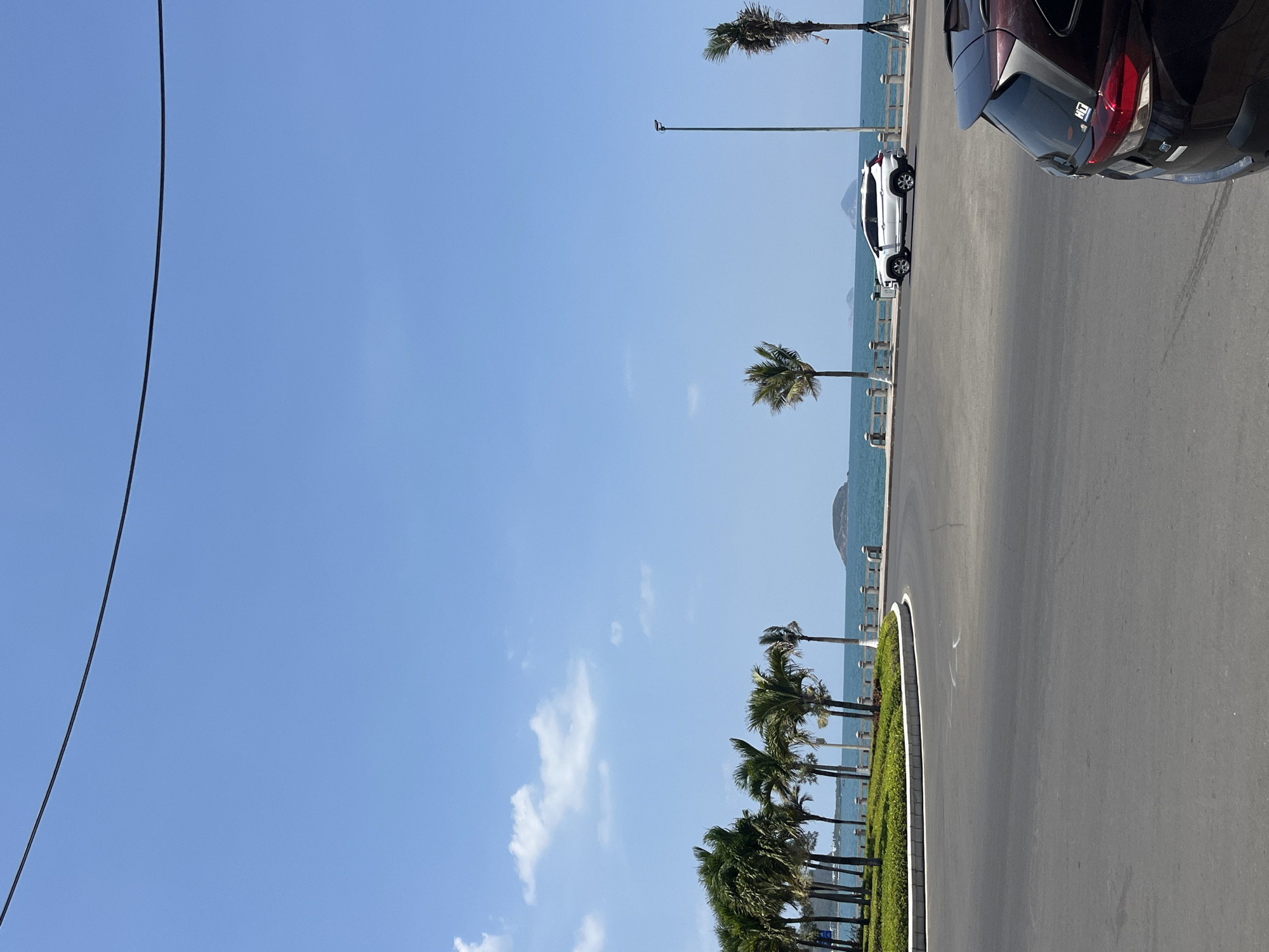 The image depicts a coastal road in Bắc Nha Trang Ward, Vietnam. A black powerline runs vertically in the upper left. The sky is clear blue with scattered clouds towards the bottom. The road runs horizontally on the right, with two parked cars partially visible. A small, green, planted median separates the road from the ocean. Palm trees line the median near the road. The ocean is visible in the background, with a small landmass or island visible in the distance. Lampposts are spaced along the median, adjacent to the water.
