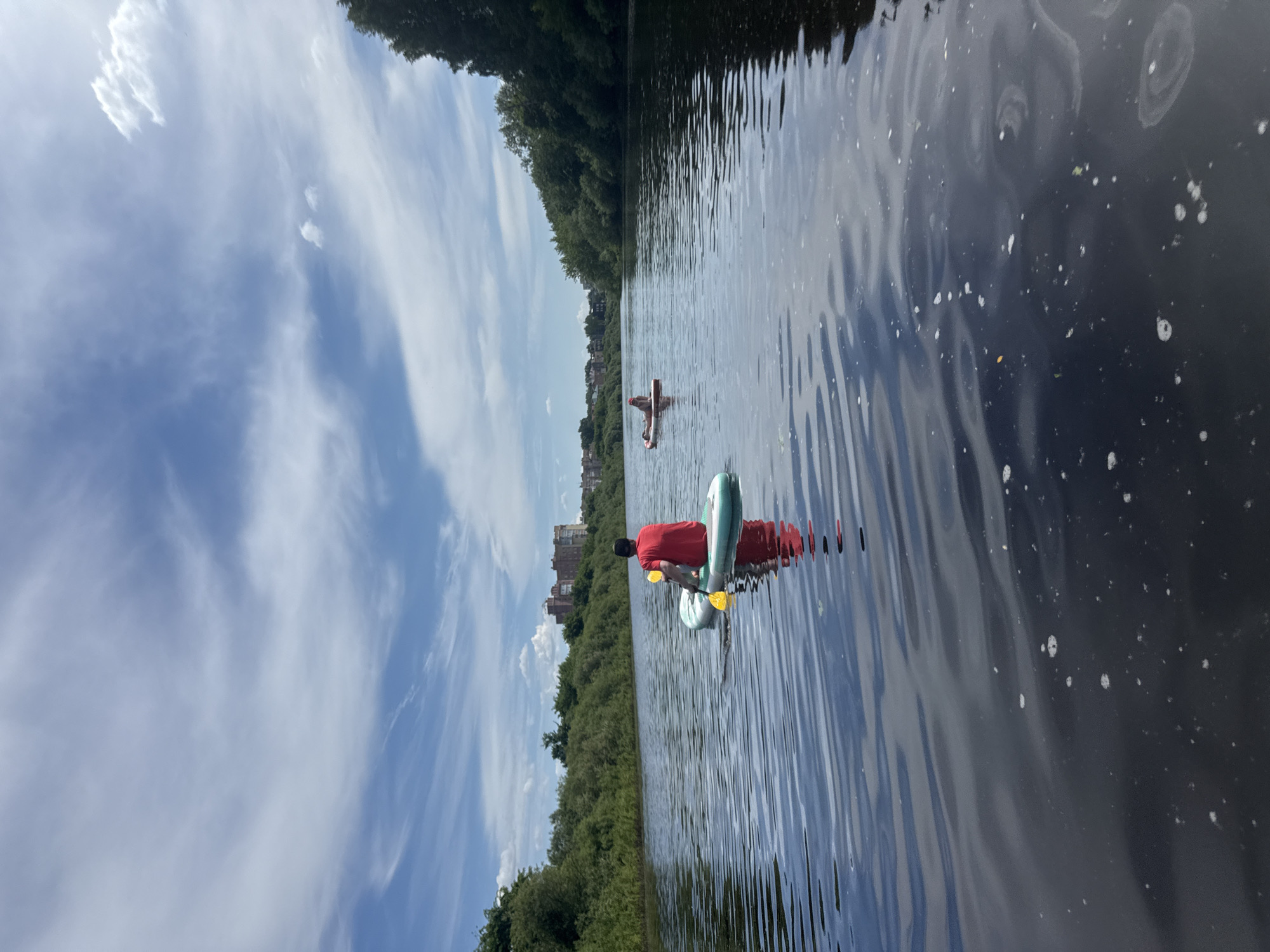 This image captures a segment of the Vologda River in Vologda, Russia, on a clear day with scattered clouds. The river's surface is dark with visible ripples, and small white foam particles are dispersed across the foreground water. A person, viewed from the back, is seated on a light teal inflatable stand-up paddleboard (SUP) in the immediate foreground. This individual wears a red short-sleeved shirt and a dark baseball cap, holding a yellow paddle in the water beside their right side. Their reflection is present on the water's surface. Further along the river, on the right side of the frame, a second individual lies prone on a red inflatable SUP. Both banks of the river are densely lined with lush green vegetation, primarily trees and bushes. In the background, on the left bank, several multi-story buildings with reddish-brown facades are visible above the treeline. The sky above is blue with distinct white, wispy clouds.