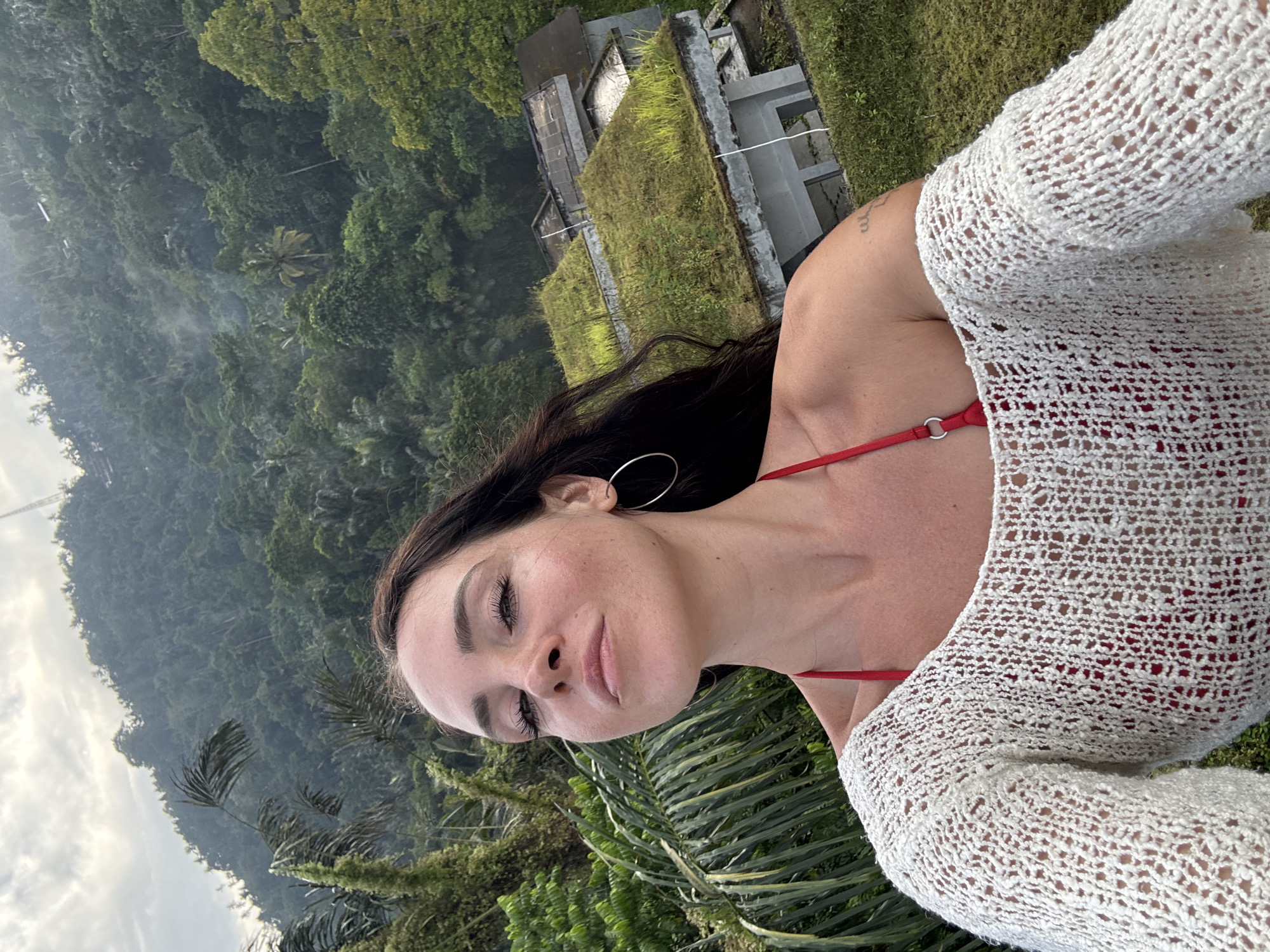 A young woman with dark hair and fair skin takes a selfie outdoors. She is wearing a red bikini top and an off-the-shoulder white knit sweater. She has large hoop earrings and appears to be looking up and to her right, with a slight smirk. Behind her, lush green jungle covers a hillside, with some buildings that have green, grass-covered roofs visible in the mid-ground. The sky is overcast with some light clouds. The scene appears to be located in Bebandem, Indonesia.