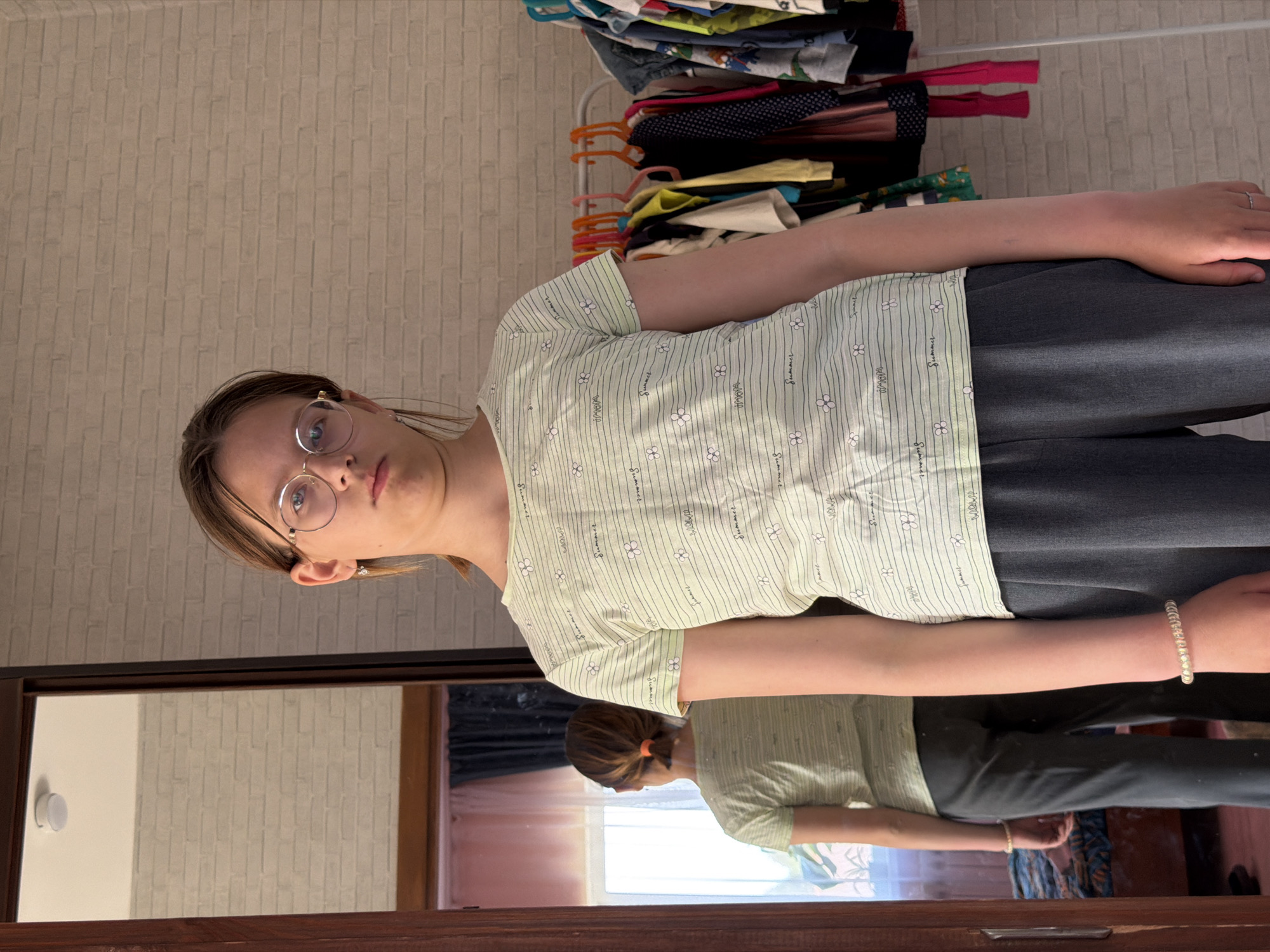 A young woman is standing in front of a mirror, with a rack of clothes visible in the background. The woman is wearing glasses, a pale green patterned t-shirt, and dark grey pants. She has a bracelet on her right wrist. The mirror reflects her image, showing her hair tied back with an orange hair tie. The background includes a wall with a brick-like pattern. The OCR in the image includes the word "Summer". The location is Chaikovsky, Russia.