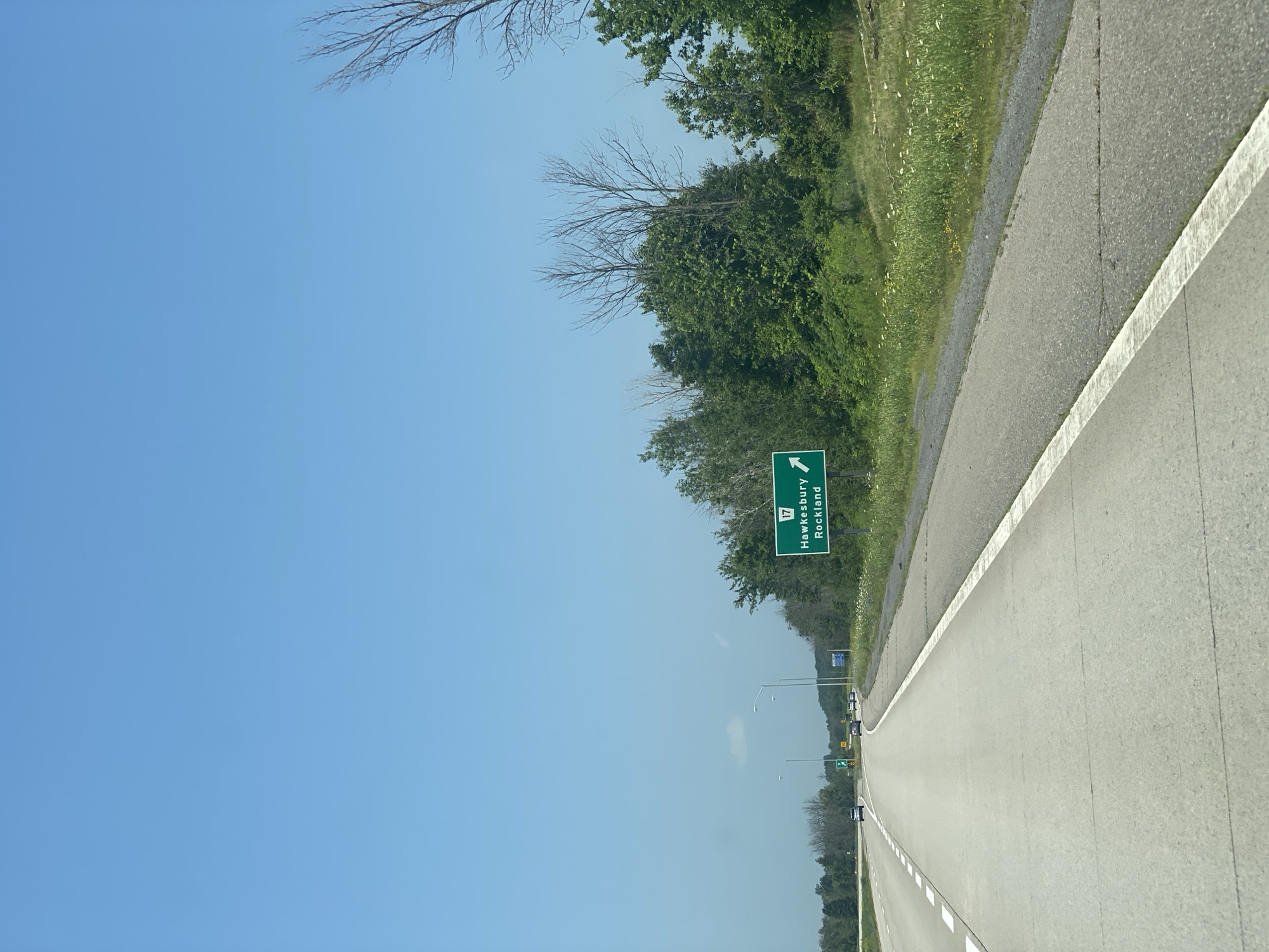 A highway sign on the right side of a highway indicates that Route 17, leading to Hawkesbury and Rockland, is to the right. The sky is clear and blue. The highway has two lanes of traffic, with a white line marking the right shoulder. There are several cars on the highway in the distance. The right side of the highway is lined with trees and green grass. The scene appears to be in East Hawkesbury, Canada.