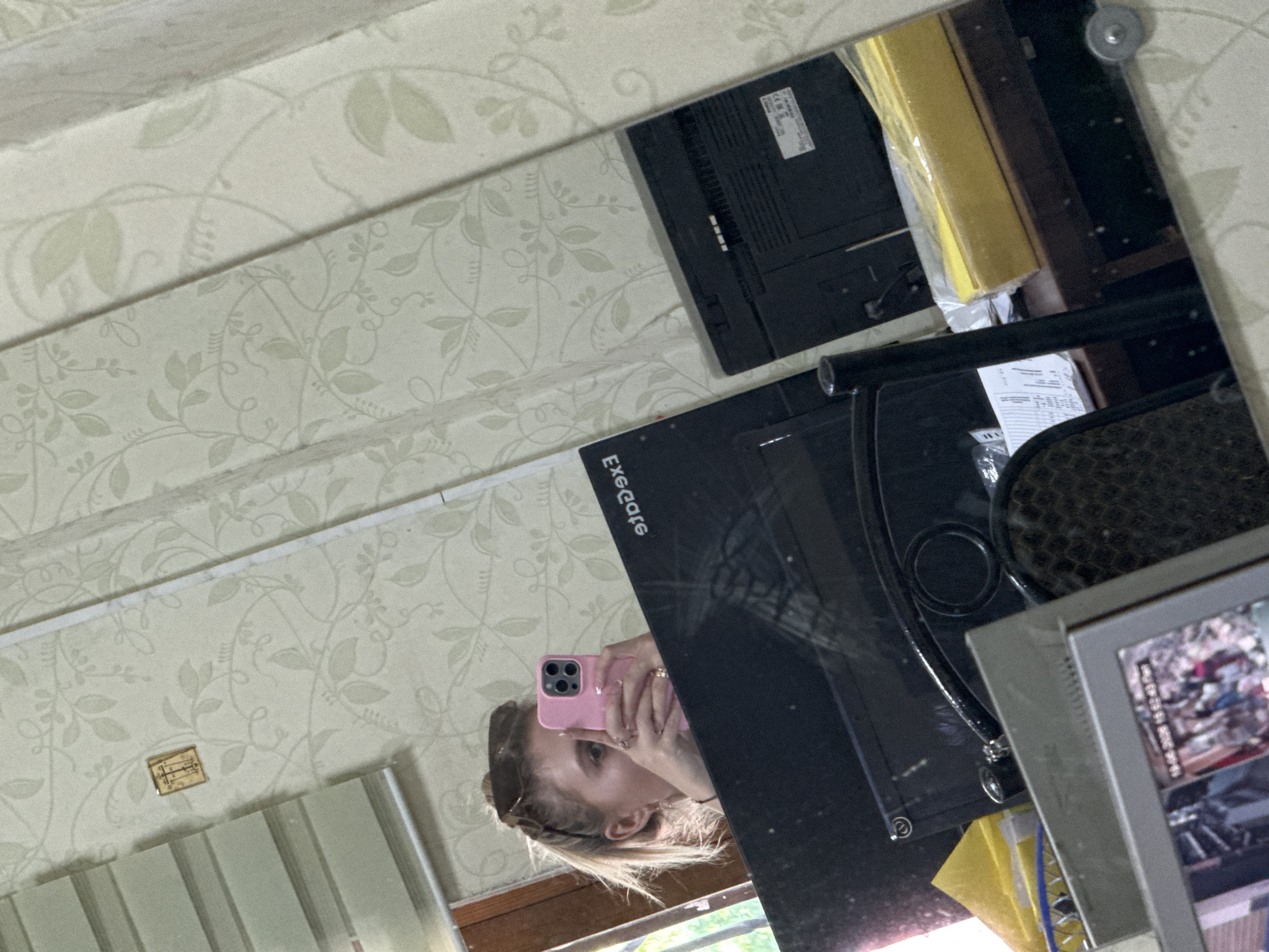 The image is a mirrored selfie of a young woman. She is holding a pink smartphone and looking at the camera. The woman has blonde hair and is wearing sunglasses. The background is a room with floral wallpaper. In the reflection, several objects are visible, including a computer monitor, a yellow item wrapped in plastic, and a framed picture. The overall impression is that the photo was taken indoors during the day. The woman appears relaxed and is smiling slightly. The visible text on the computer monitor appears to be in English, possibly a brand name or model number, but it is not entirely clear. The framed picture contains a photograph of people, possibly a family.