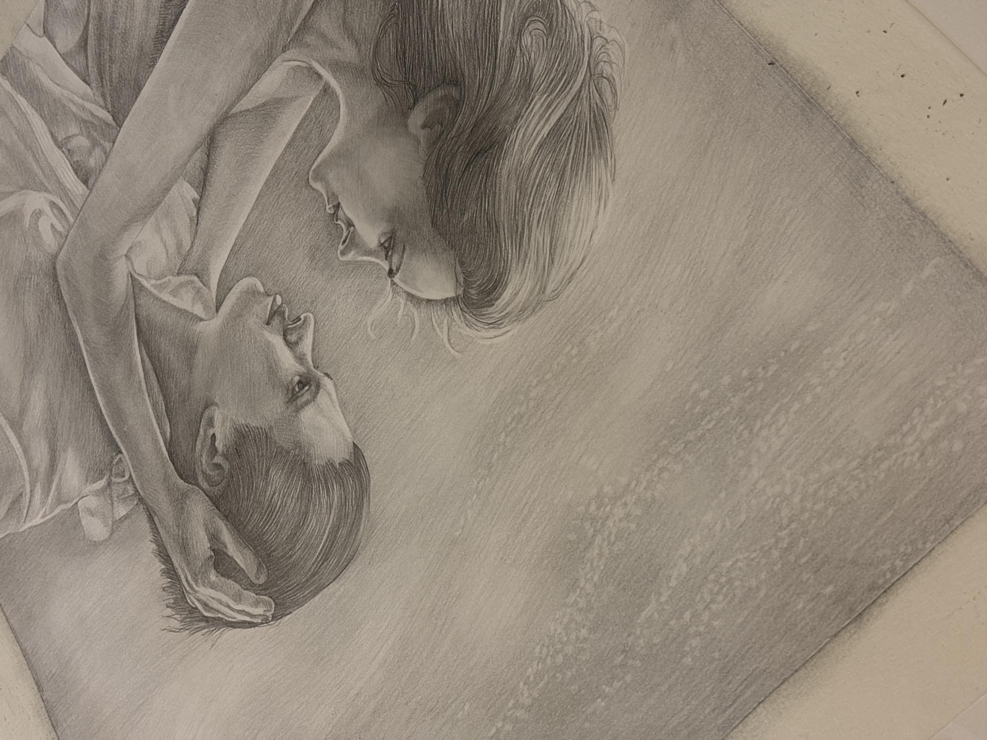 This monochrome pencil drawing depicts two individuals, likely a man and a woman, in an intimate and tender embrace. They are shown lying down, with the woman positioned above and gazing down at the man, whose eyes are directed upwards towards her. Their faces are very close, conveying a strong sense of affection, connection, and peace.
The woman has long, flowing hair, and a soft, gentle smile adorns her face. Her arm is draped across the man's body. The man's expression appears serene or adoring, and one of his hands is gently resting near his head, possibly supporting it or caressing his own hair. Both figures are rendered with noticeable detail, showing realistic features, hair textures, and the folds in their light-colored clothing.
The artwork is characterized by its skillful use of pencil shading, which creates depth, form, and a lifelike quality. The background is softly shaded, textured with visible pencil strokes, and appears to be a blanket or a soft surface, contributing to the intimate setting. The composition places the figures diagonally across the left side of the frame, leaving a significant portion of the right side as a lightly shaded, textured background, which emphasizes the interaction between the subjects. There is no visible text in the image. The overall mood is one of quiet intimacy and emotional warmth.