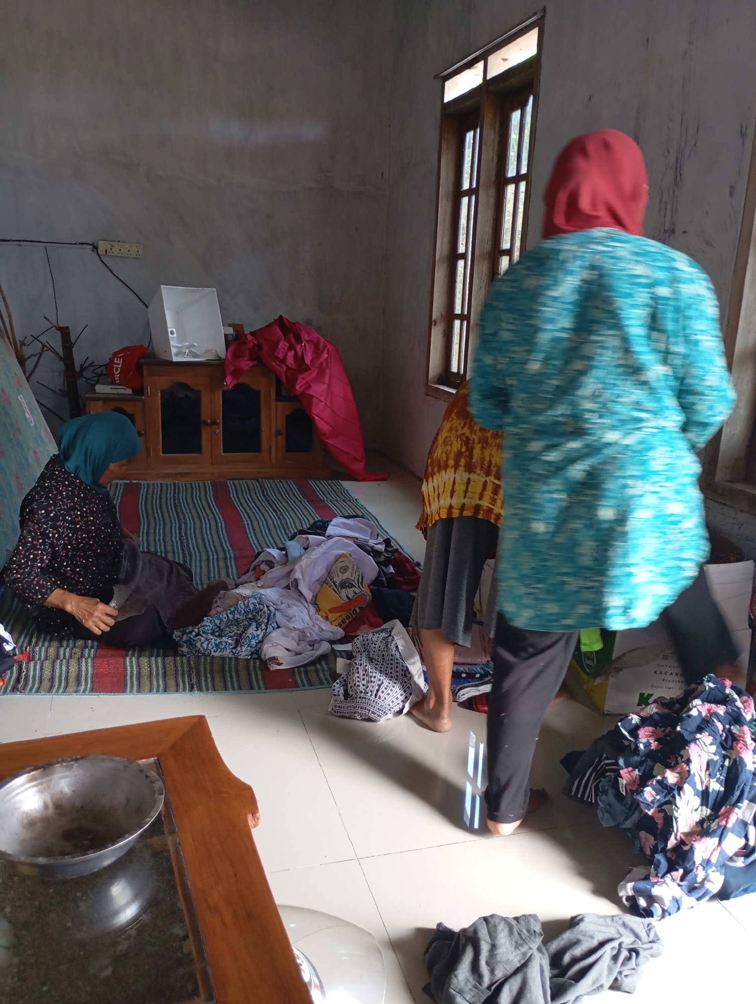 In a dimly lit room with bare concrete walls, two women are engaged in domestic chores. One woman, wearing a floral-patterned dress and a green headscarf, sits on a striped mat and appears to be folding or sorting laundry. A pile of colorful clothes is spread out in front of her. Another woman, wearing a turquoise shirt and a red headscarf, stands with her back to the camera, also near a pile of clothes. A wooden cabinet with glass doors stands against the wall, and a red piece of fabric is draped over it. A window with wooden panes allows natural light into the room, illuminating some of the clutter. A wooden table with a metal bowl on it is visible in the foreground. The scene suggests a humble living space in Ketepung, Indonesia, where everyday activities are taking place.