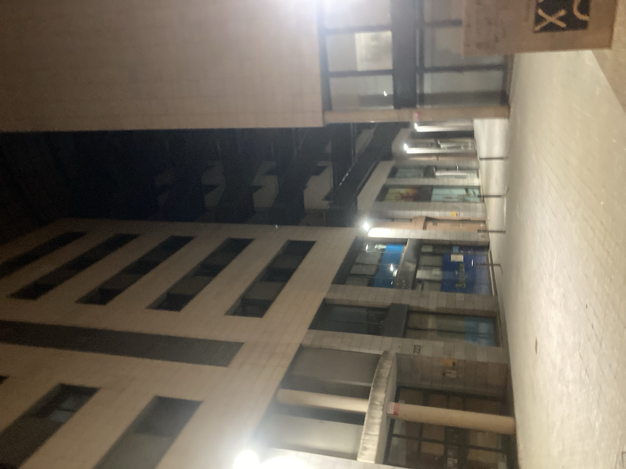 The image shows a dimly lit, modern street at night, likely in a commercial or residential area of Barcelona, Spain. Two multi-story buildings flank a pedestrian walkway paved with light-colored tiles. The building on the left has a light-colored facade with a regular pattern of dark horizontal bands and recessed windows. The building on the right has a lighter, smoother facade with large glass windows on the ground floor. The walkway is lined with bollards, and streetlights are visible, casting a warm glow on the scene. The overall atmosphere is quiet and somewhat stark, suggesting an urban environment after business hours. No people are visible in the frame. The cropped images provide close-ups of the building facades, windows, and tiling.