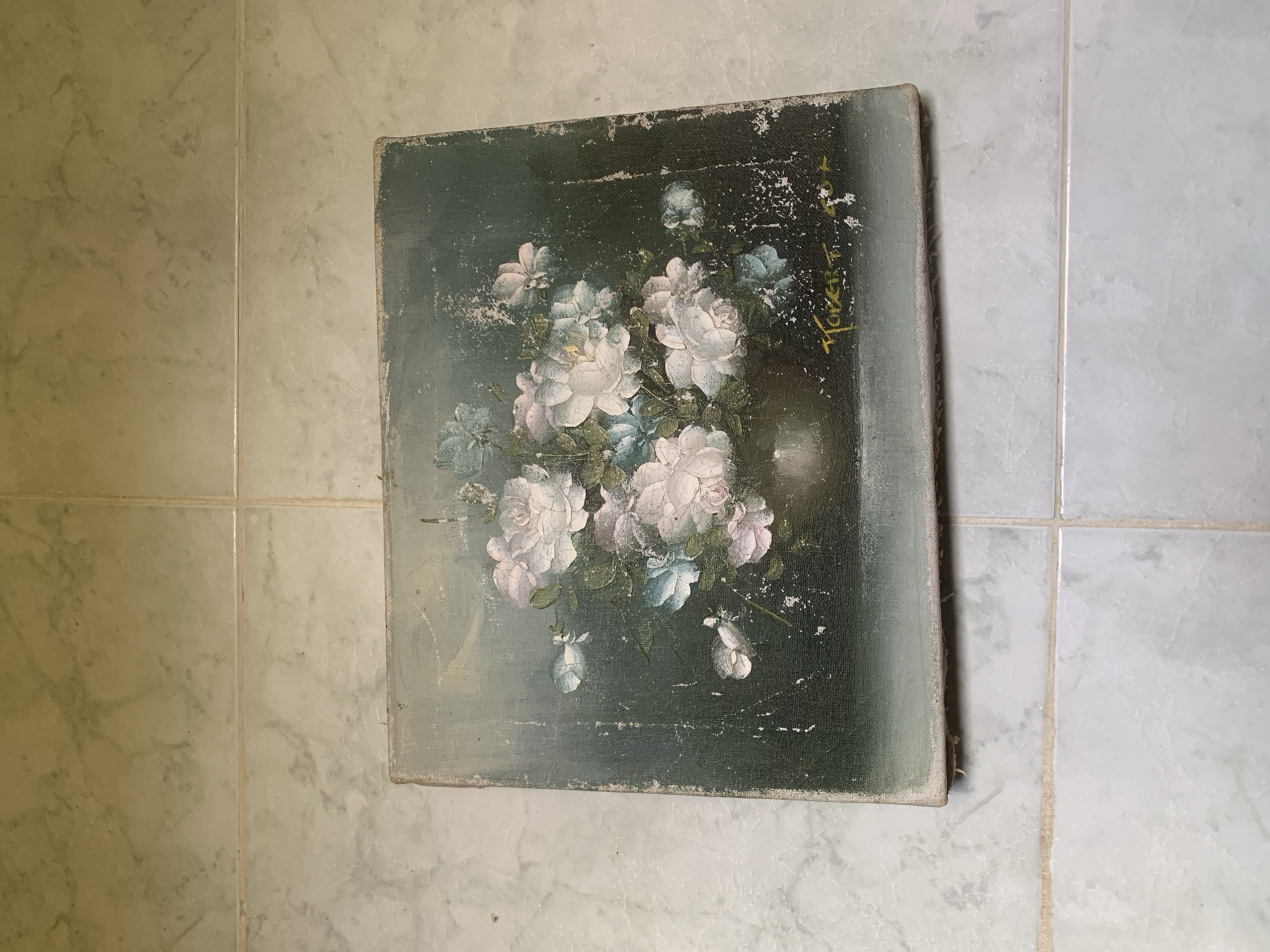A square, somewhat deteriorated painting of a bouquet of roses rests on a floor covered in square, light-colored tiles. The painting features a dark green and bluish-grey background with a cluster of white and light pink roses in the center. There is a signature visible in the upper right corner, appearing to read "Flores T. 1944." The painting's edges are worn, and the tiles have a mottled texture, suggesting age or wear. The location cannot be determined to be Santa Ana, Venezuela based on the visual content of the image.