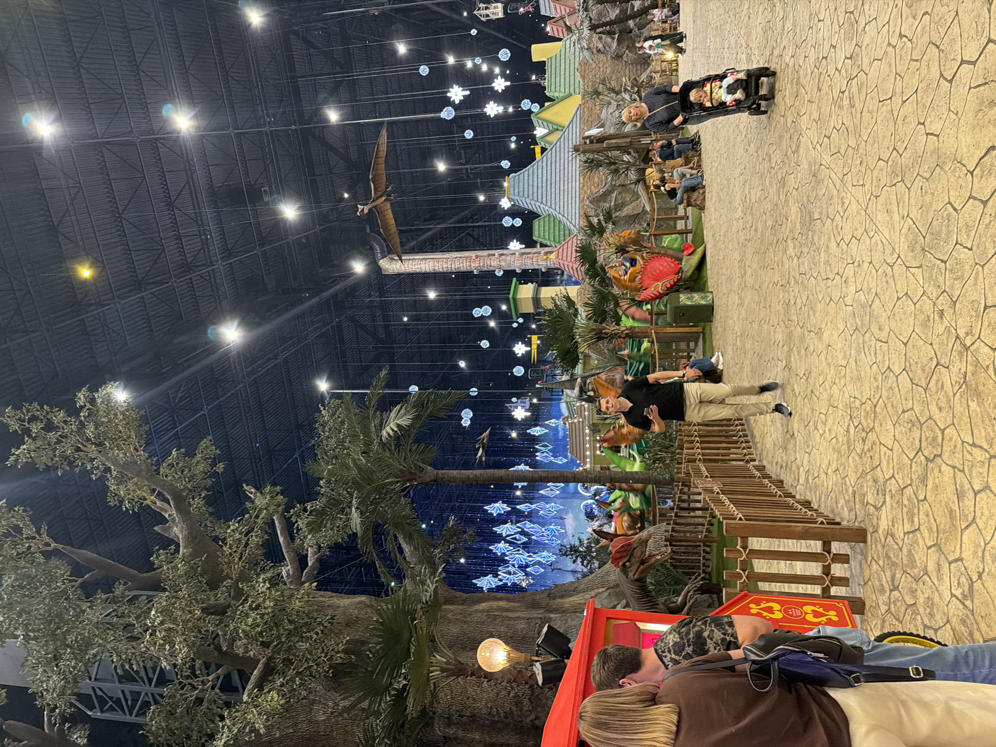 The image depicts an indoor scene, likely an amusement park or entertainment venue, with a dinosaur theme. In the foreground, several people are visible. A man in a black shirt and beige pants stands on a stone-like walkway. To his right, a wooden bridge-like structure is visible. In the lower part of the image, two people are lying down, one with a camouflage-patterned shirt, and a blue bag is resting on them. A bright yellow light bulb is also present on the left.
The background reveals a large, cavernous space with a high, dark ceiling crisscrossed by metal beams and lit by numerous lights. Hanging from the ceiling are decorative elements, including what appear to be star or snowflake-shaped lights in blue and white, and a large model of a pterodactyl suspended in mid-air. Further back, there are structures resembling small buildings with colorful roofs, palm trees, and other dinosaur models, including a red dinosaur figure. A stroller with two young children is pushed by a woman wearing a polka-dot dress. Other people are seated and standing around the area, suggesting a public gathering.
The lighting suggests it is likely evening or indoors with artificial illumination. The overall atmosphere seems relaxed and family-oriented, with elements of entertainment and recreation. There is no visible text in the image.