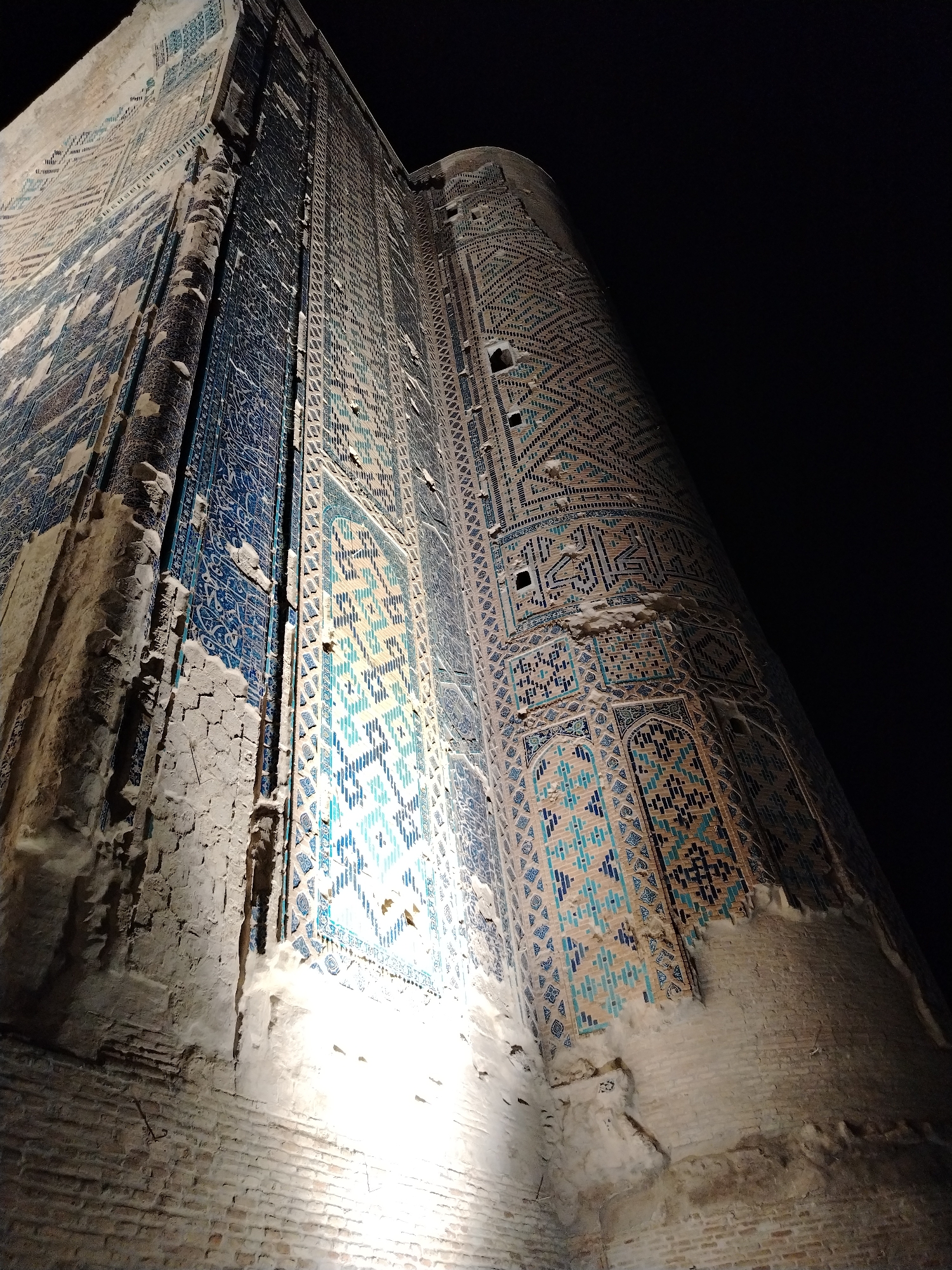The media file displays a low-angle, wide shot of a large, ornate building, possibly a mausoleum or mosque, at night in Shahrisabz City, Uzbekistan. The building's facade is covered in intricate geometric tilework, primarily in shades of blue, turquoise, and white. The tiles form complex patterns, including repeating diamond shapes and what appear to be Arabic inscriptions.
The structure is massive, with visible brickwork at its base, some of which is crumbling and aged. Large sections of the tilework have fallen away, revealing the underlying brick and plaster, giving the building a weathered and ancient appearance. The contrast between the illuminated lower portions and the dark night sky creates a dramatic effect. Artificial lighting illuminates the bottom part of the structure, casting bright lights upwards and highlighting the textures of the brick and the lower tiles.
The perspective from below emphasizes the towering height and grandeur of the building. The scene is static, with no people or activity evident. The time of day is clearly night due to the black sky and artificial lighting. The weather is not discernible. The architectural style and the presence of elaborate tilework are strong indicators of Central Asian or Islamic architecture. There is no visible text that can be clearly read or deciphered from the image.
