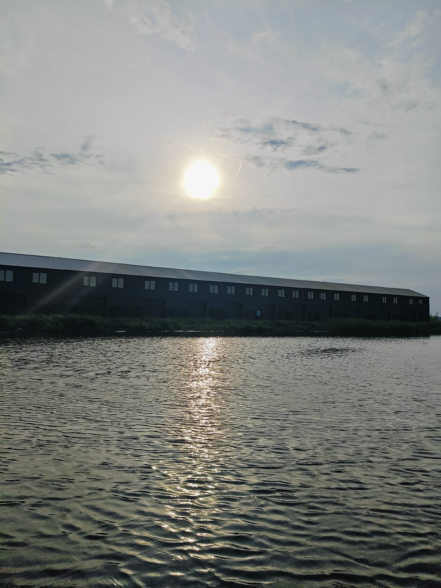 The image depicts a long, dark building with a metal roof, featuring multiple uniformly spaced windows along its facade. The building is situated on a grassy embankment next to a body of water. The water's surface is rippled and reflects the sunlight, creating a shimmering effect. In the upper portion of the image, the sun is visible in a hazy sky with scattered clouds. The scene appears to be a landscape photograph taken from a boat or the edge of the water, looking towards the building. The location is identified as being in the city of Chaikovsky, Russia. The image is devoid of people or any explicit signs of human activity beyond the structure itself.