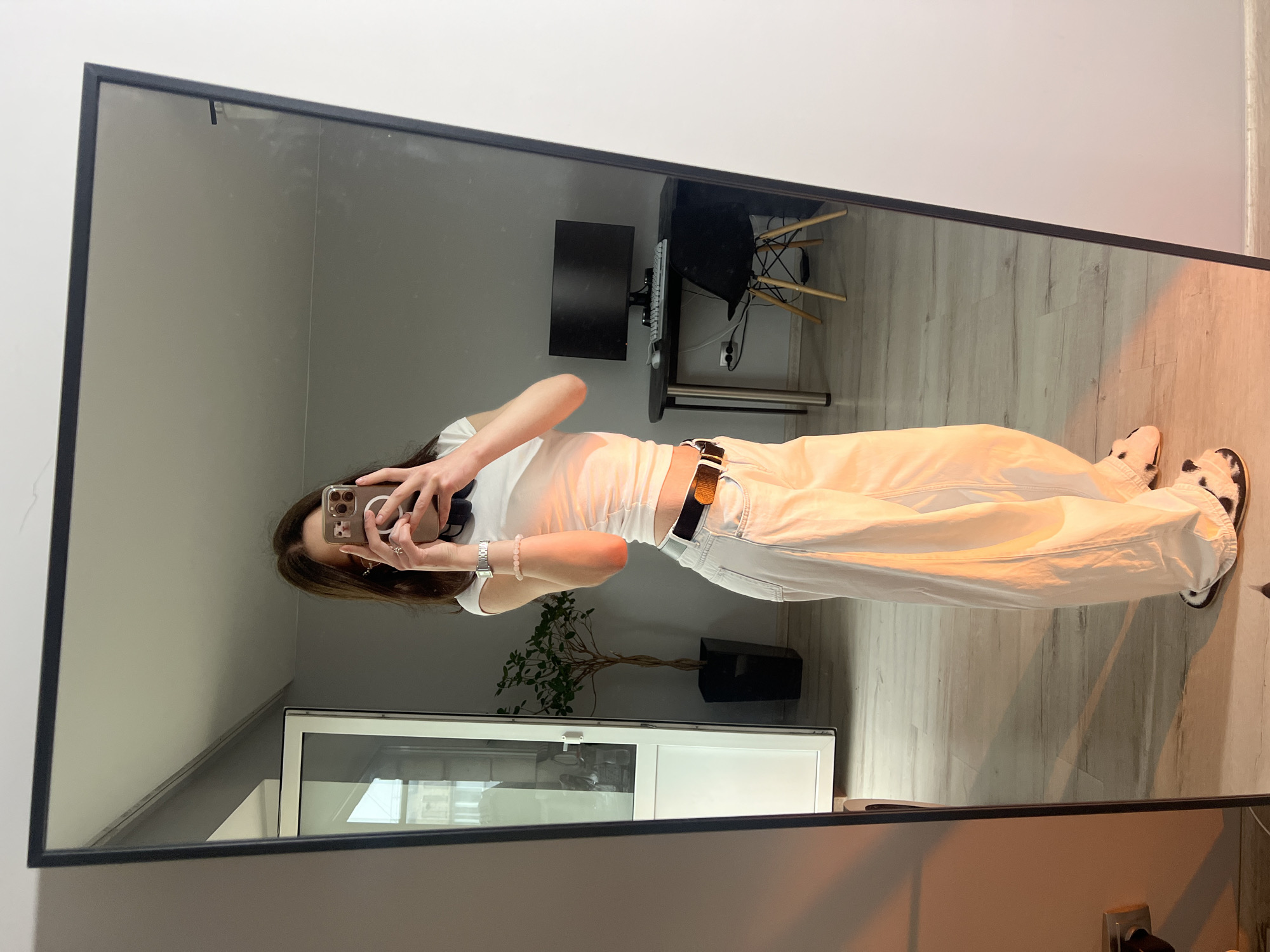A person is captured in a mirror selfie, standing upright and holding a smartphone with both hands to take the picture. The individual, who appears to be female with long brown hair, is wearing a white short-sleeved top, light-colored baggy pants, and a dark belt with a silver buckle. A silver watch and a light pink beaded bracelet are visible on her left wrist. She is also wearing black and white patterned shoes.
The scene reflects an indoor setting, likely a room or office, with light gray walls and light-colored wooden or laminate flooring. In the background of the reflection, a black desk is visible with a computer monitor, keyboard, and a black office chair featuring wooden legs. A plant in a black rectangular pot sits on the floor near a white cabinet with glass panels. A power outlet is also discernible near the desk. The room is well-lit, with a warm-toned light casting a glow on the lower part of the person's body and the floor, possibly from an artificial light source or angled sunlight. There are no legible texts visible. The location context for this media file is Ternopil, Ukraine.