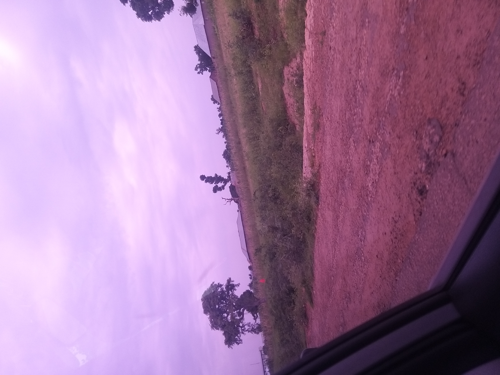 The image shows a landscape scene with a dirt road in the foreground and a gentle hill in the background. The road is made of reddish-brown gravel and appears to be uneven, with some potholes visible. The hill is covered with green vegetation and some scattered trees. In the background, there are a few buildings with pitched roofs, suggesting a rural or semi-urban area. The sky is a pale purple, indicating either dawn, dusk, or possibly an effect of lighting conditions. A part of a vehicle's interior, likely a car window frame, is visible in the lower left corner of the image, implying the photograph was taken from inside a vehicle. There are no people or animals clearly visible in the scene. The overall impression is that of a quiet, undeveloped area. Without specific landmarks or distinguishing features that are unique to Dukku, Nigeria, it is not possible to confirm the location within the city based solely on this image.