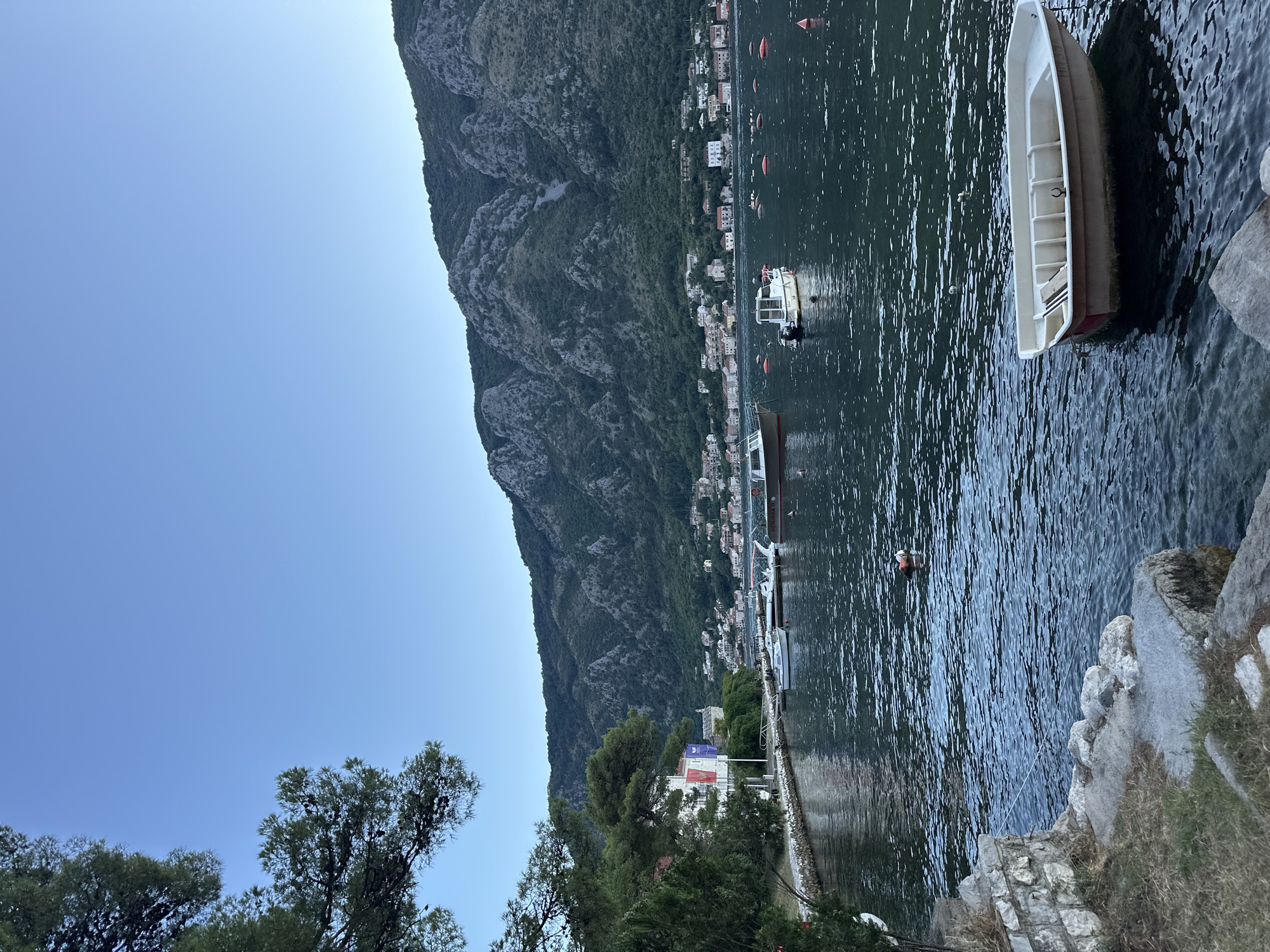 The image captures a serene waterfront scene in Dobrota, Montenegro, likely during late afternoon or early evening given the soft light and clear sky. The foreground features a stone embankment with dry grass and rocks leading into the calm, dark water. Several small boats are present: a white rowboat is moored in the immediate foreground, its interior visible. Further out in the bay, other boats, including a larger motorboat and several smaller vessels, are anchored or tied to buoys. The water is slightly rippled, reflecting the surrounding landscape.
In the midground, a waterfront promenade is lined with a stone wall and lush green trees. Buildings with red-tiled roofs, characteristic of coastal Mediterranean architecture, are visible along the shore, nestled against a backdrop of steep, verdant mountains. The mountains are densely covered with trees, with rocky outcrops interspersed. The sky is a clear, pale blue, transitioning to a slightly darker hue near the horizon, suggesting the sun is beginning to set or has recently risen. There are no discernible people in the immediate foreground or midground. Small orange buoys dot the water, marking channels or mooring areas. The overall atmosphere is one of tranquility and natural beauty.