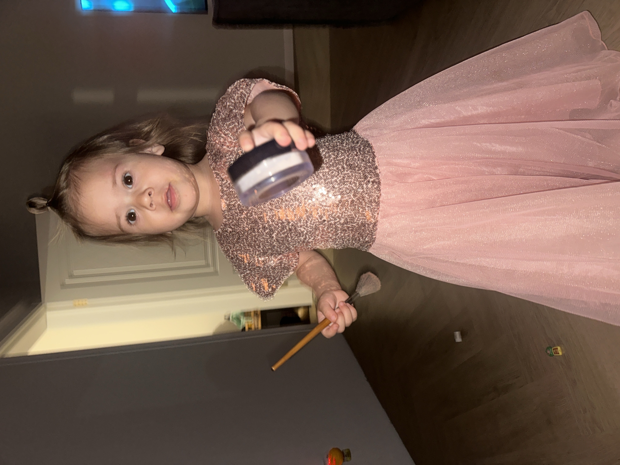A young girl wearing a pink sequined dress is holding a makeup brush and a container of powder. The scene is indoors. Kharnavy, Russia is not mentioned in the image, so the location cannot be identified.