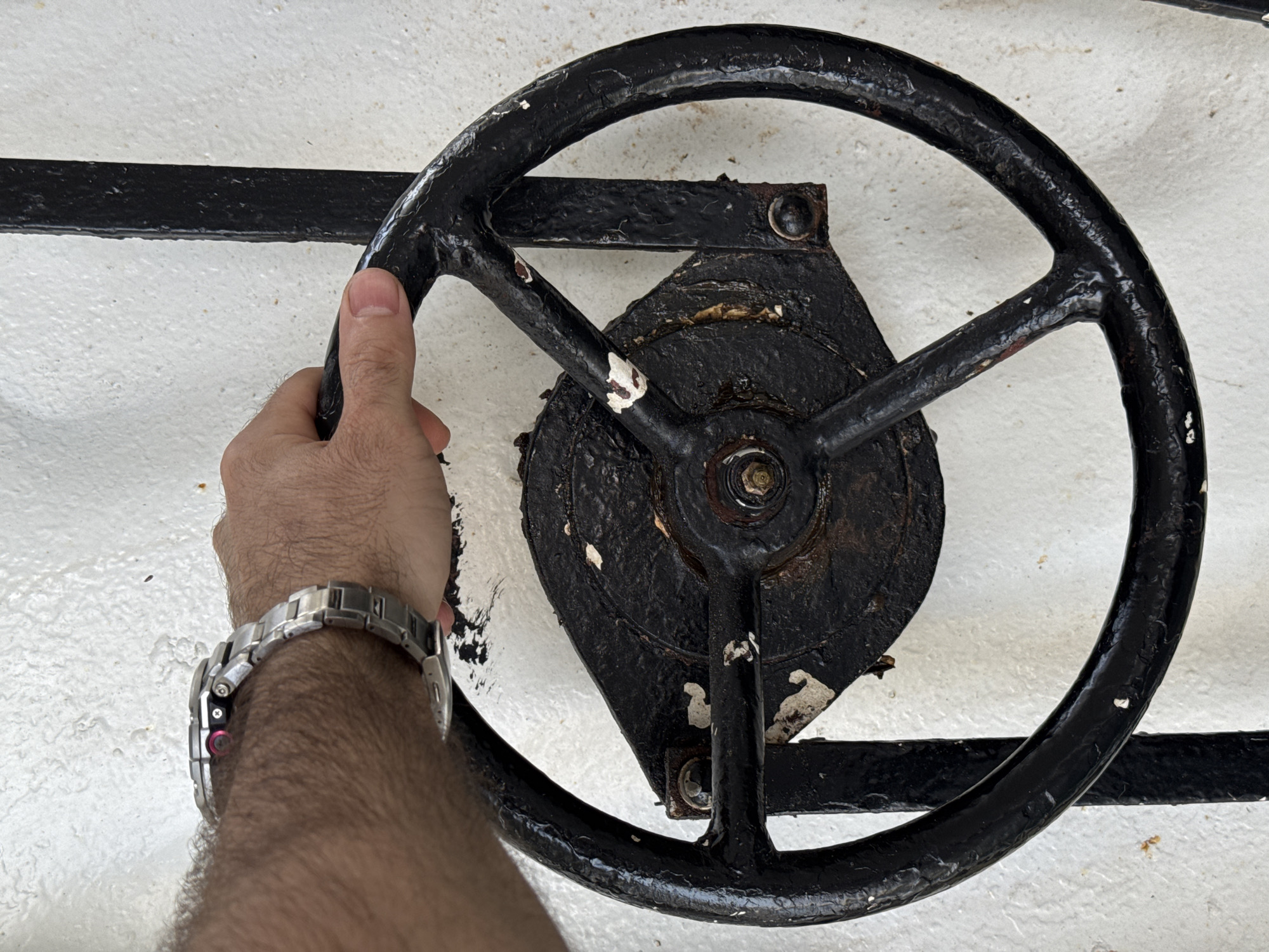 A hand with a silver wristwatch is holding a black steering wheel. The steering wheel is attached to a black mechanism that is bolted to a white surface. There is a black horizontal bar behind the steering wheel. The steering wheel is worn and has some chipped white paint. The background is a textured white surface. The lighting suggests it is daytime, but the specific time of day is not discernible. There are no visible landmarks or environmental cues to determine the location or weather. The image appears to be a close-up shot focusing on the hand and the steering wheel. There is no visible text in the image.