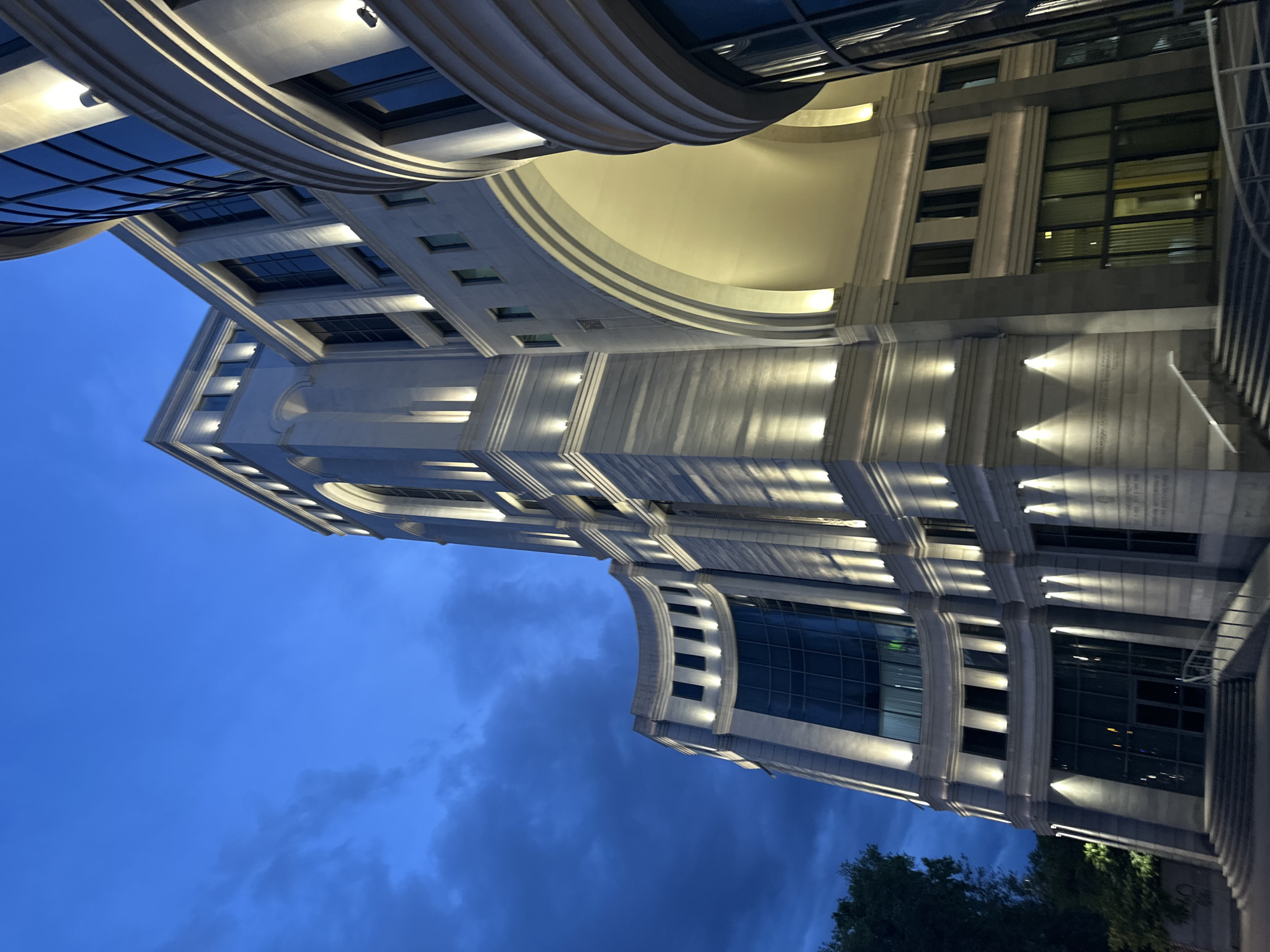 The image shows a modern architectural building at night, likely located in Yerevan, Armenia. The building features a combination of glass and stone facade, with lit-up architectural details accentuating its structure. The sky above shows a deep blue hue, suggesting either twilight or a night scene with artificial illumination. Parts of trees are visible at the bottom of the frame.