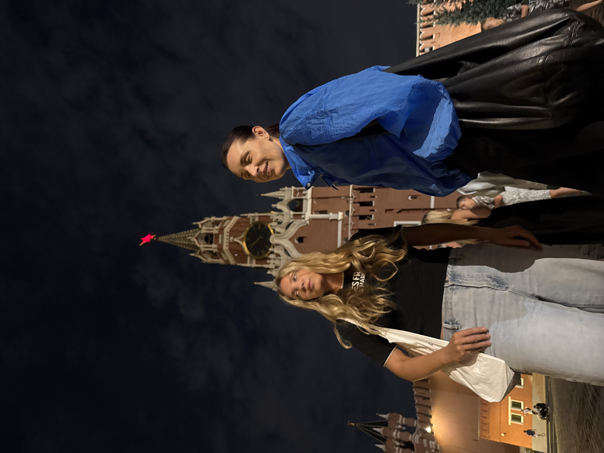 The image captures a nighttime scene in Red Square, Moscow, Russia, with the iconic Spasskaya Tower prominently featured. Two women are in the foreground, positioned in front of the tower. The woman on the left has long blonde hair and is wearing a black t-shirt and light-wash wide-leg jeans. She carries a white tote bag over her shoulder. The woman on the right, with dark hair pulled back, is wearing a bright blue windbreaker over a dark garment, and a large black leather bag is slung over her shoulder.
The Spasskaya Tower, a prominent landmark of the Moscow Kremlin, is illuminated against the dark night sky, with its clock face visible and a red star atop its spire. The surrounding Kremlin wall is also visible, with some warm lighting from within the complex. The sky is dark with some visible clouds. In the background, other people can be seen in the distance, some sitting and others standing, suggesting a public gathering or pedestrian activity within the square. The overall atmosphere is one of urban exploration at night, with architectural grandeur as a backdrop.