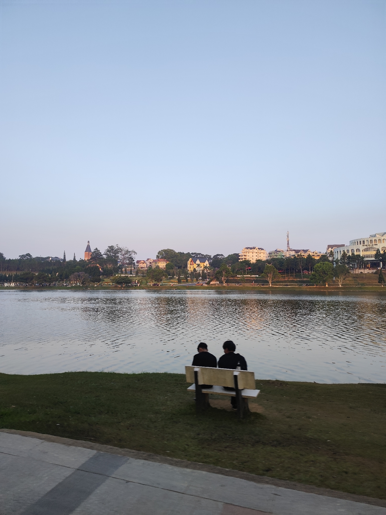 The image shows two people sitting on a bench overlooking a lake in Da Lat, Vietnam. The location is near Xuan Huong Lake. The lake is in the middle ground, with the city skyline visible in the background, including notable buildings and trees. The people are seated on a white bench on a grassy area. In the foreground, a portion of a concrete walkway is visible. The sky is pale blue. The water has visible ripples.