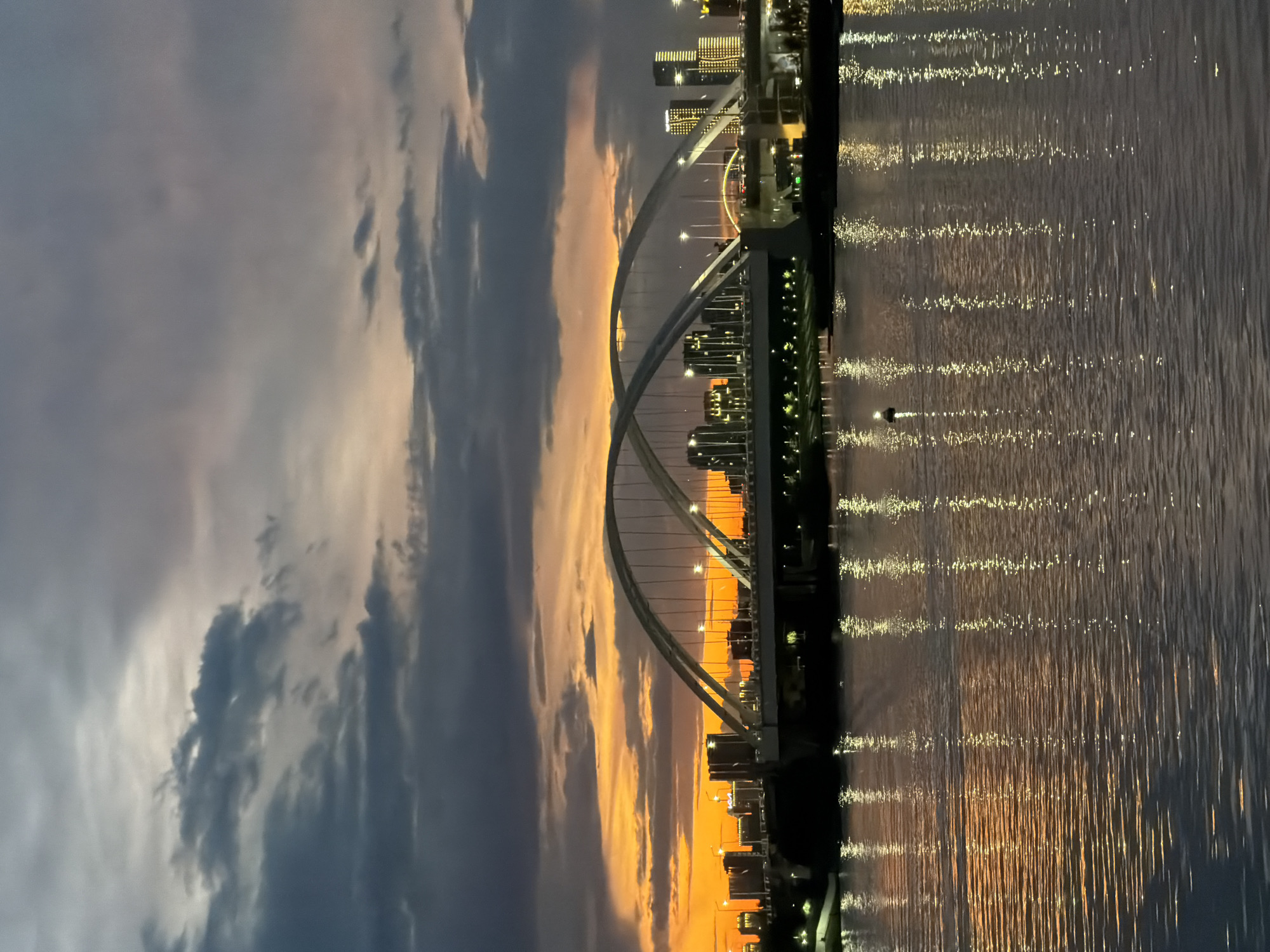 The image captures a serene twilight scene of the Bayterek bridge in Astana, Kazakhstan. The bridge, a prominent architectural landmark, features a distinctive arched design, with its lighting system illuminated against the darkening sky. The vibrant hues of the sunset, with shades of orange and yellow, streak across the horizon, contrasting with the deep blues and grays of the clouds. The calm waters of the Ishim River reflect the city lights and the vibrant sky, creating a shimmering, textured surface.
The scene depicts a tranquil moment, likely at dusk, as the city transitions from day to night. The lights of the buildings along the riverbank are starting to twinkle, and the bridge itself is a beacon of light. The mood is peaceful and contemplative, evoked by the soft glow of the sunset and the gentle ripple of the water. There are no visible people in the immediate foreground, adding to the sense of solitude and the vastness of the landscape. The image is a beautiful representation of Astana's urban landscape merging with natural beauty at the end of the day.
