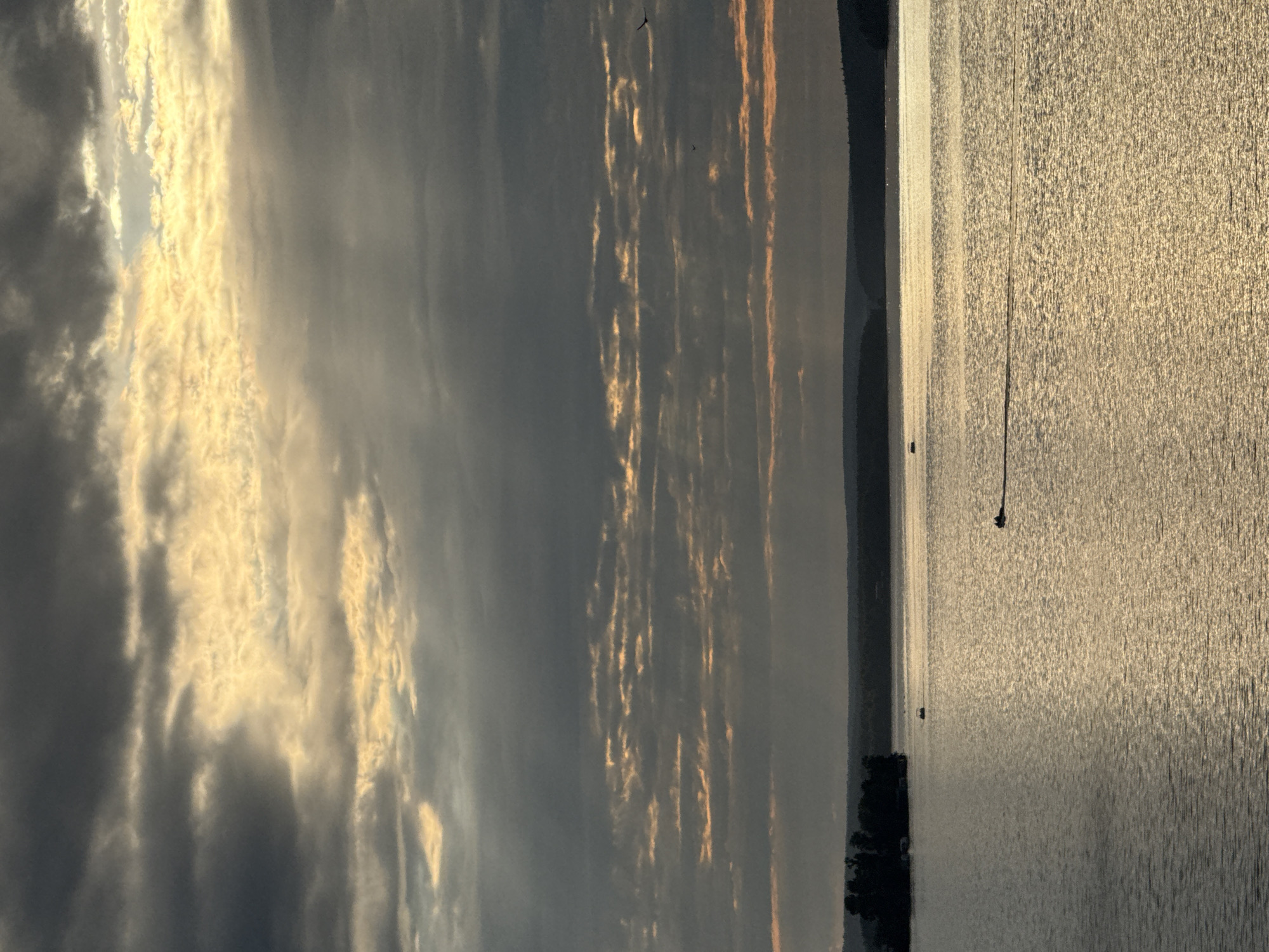 A wide-angle, eye-level shot captures a body of water reflecting golden light from the sky. The sky is filled with dramatic, layered clouds in shades of grey and yellow, with some areas illuminated by sunlight breaking through. A dark, forested shoreline with rolling hills is visible in the distance. On the water, a small boat with two occupants is moving from right to left, leaving a wake. Another small boat is visible further in the distance to the left. A small, dark, tree-covered island is situated in the upper left quadrant of the image. The overall mood is serene and atmospheric.