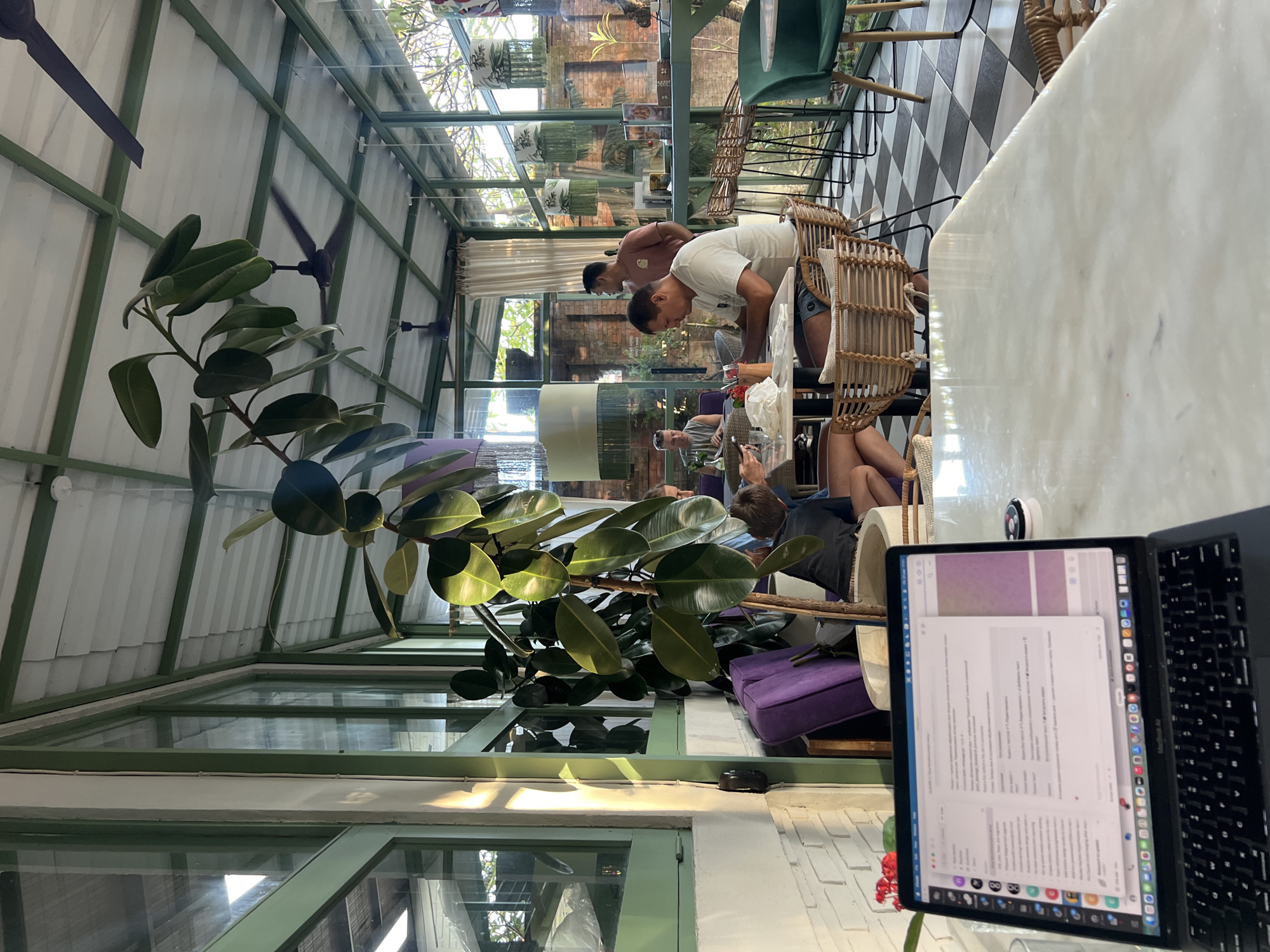 The photograph captures an interior scene in Sukawati, Indonesia, featuring a glass-enclosed space with a geometric metal frame. Inside, patrons are seated at tables, some engaging with laptops, while others converse. The lighting is bright, with natural light filtering through the glass walls. Large-leaved plants are interspersed throughout the seating area, adding a touch of greenery. The flooring is a black and white checkered pattern. The scene suggests a café or co-working space environment.