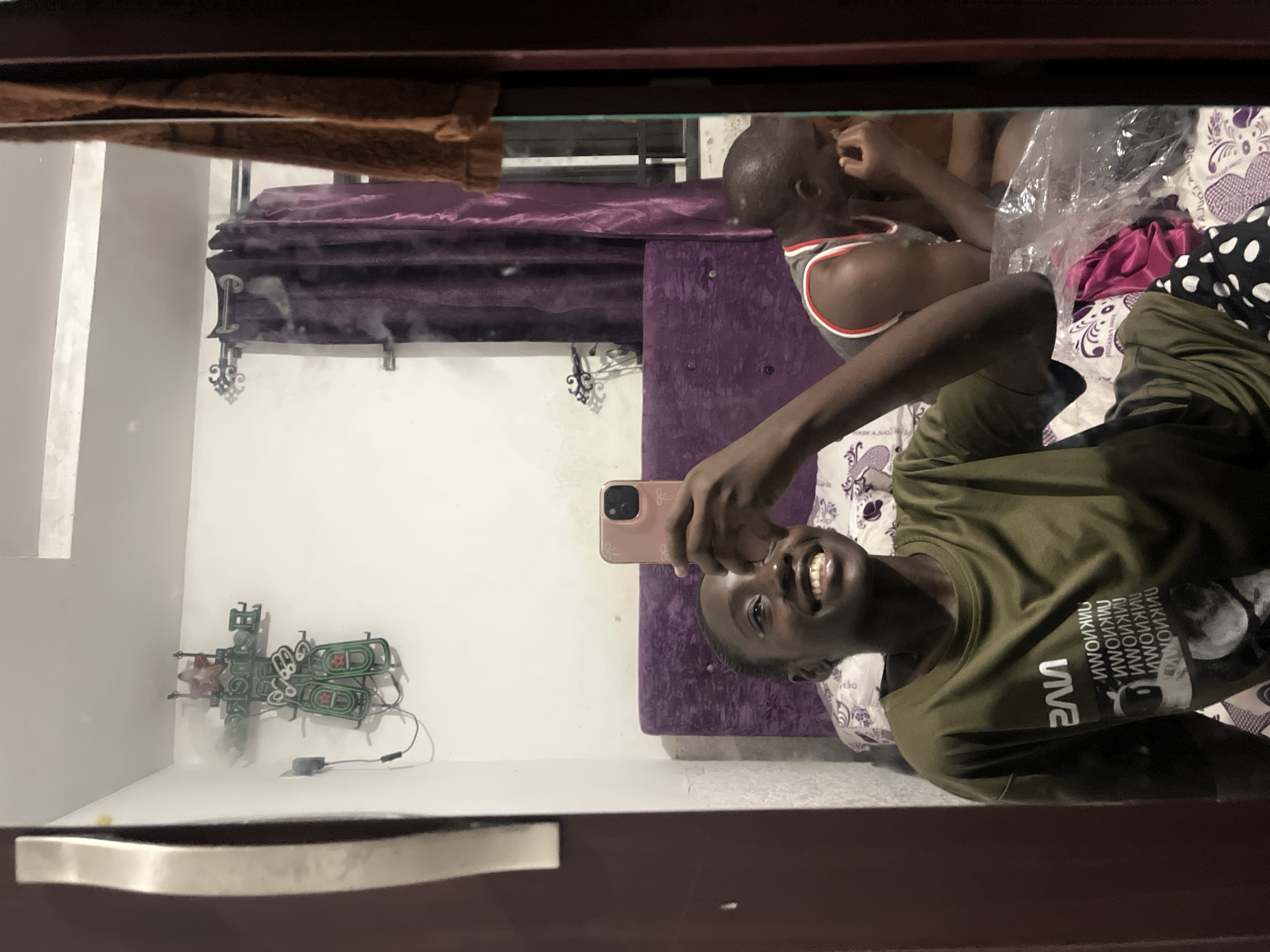 The image is a selfie taken in a mirror within a room. Two individuals are visible: the person taking the selfie and another person lying on a bed in the background. The person taking the selfie is dark-skinned, wearing an olive-green t-shirt, and smiling. The second individual is lying on the bed, wearing a tank top.
The room contains a bed with purple headboard, pillows with patterned cases, and blankets. A neon sign is mounted on the wall. The mirror is framed with a dark-colored frame.
There is no identifiable information in the picture that suggests that the location is Abuja, Nigeria.