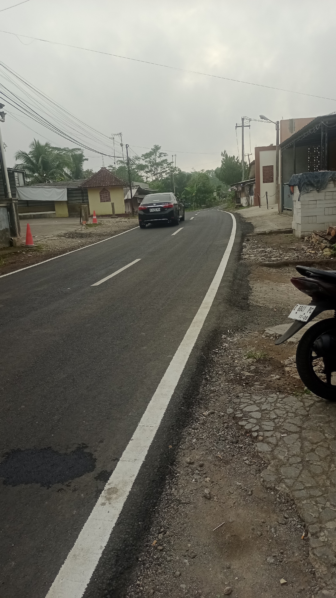 A black sedan with license plate number P 1472 ALL is driving on a paved road in Cogreg, Indonesia. The road has a white dividing line and is surrounded by houses and vegetation. The sky is overcast. A motorcycle is parked on the side of the road.