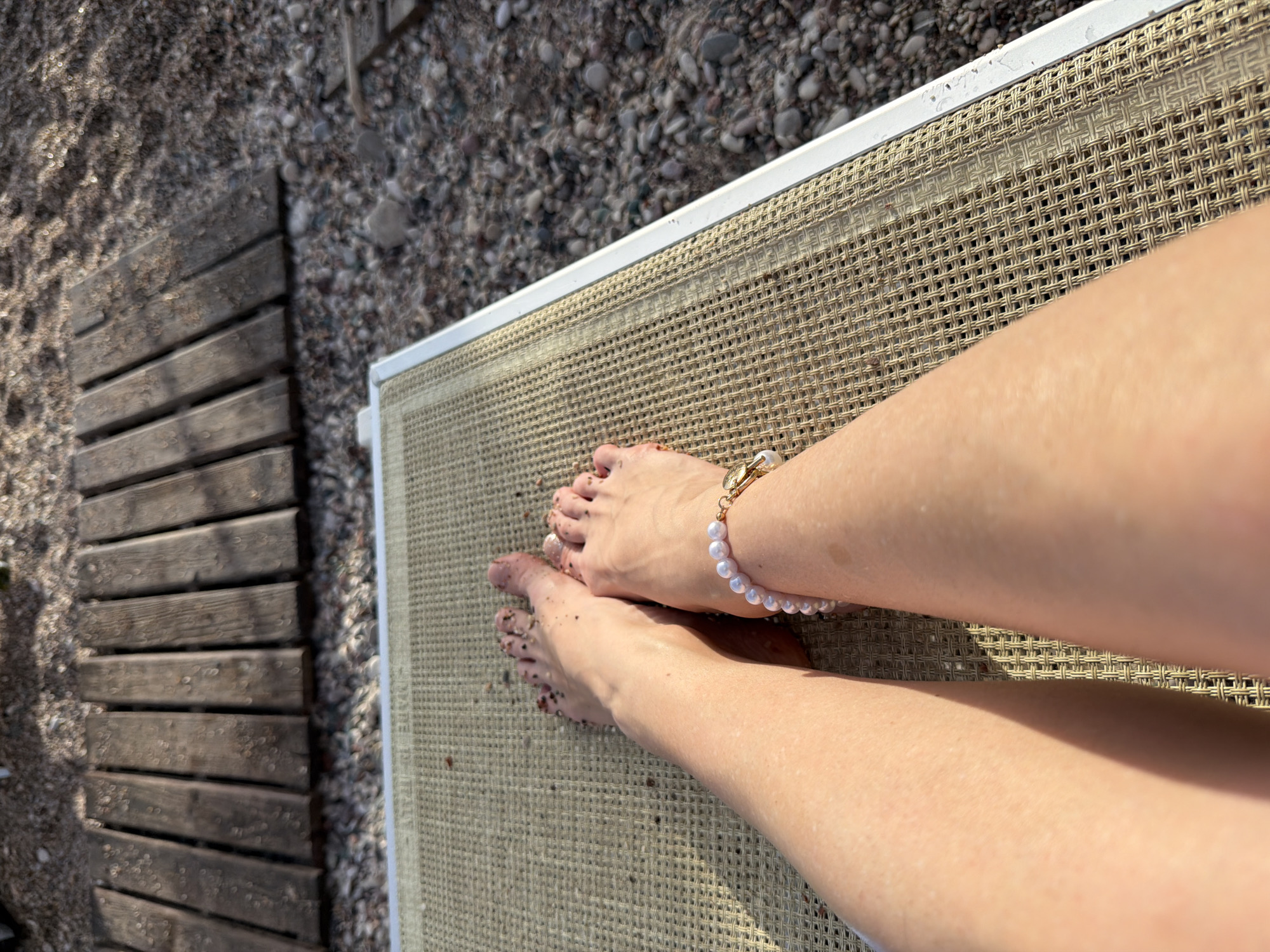 The image shows a close-up view of a person's legs and feet resting on a beige mesh beach lounger. The lounger is positioned on a pebbly beach, with a wooden walkway visible in the background. The person's feet are bare and appear to have sand on them. A pearl bracelet with a gold charm is worn on the left ankle. The scene is likely in Kamenovo, Montenegro, given the typical landscape of the area.
