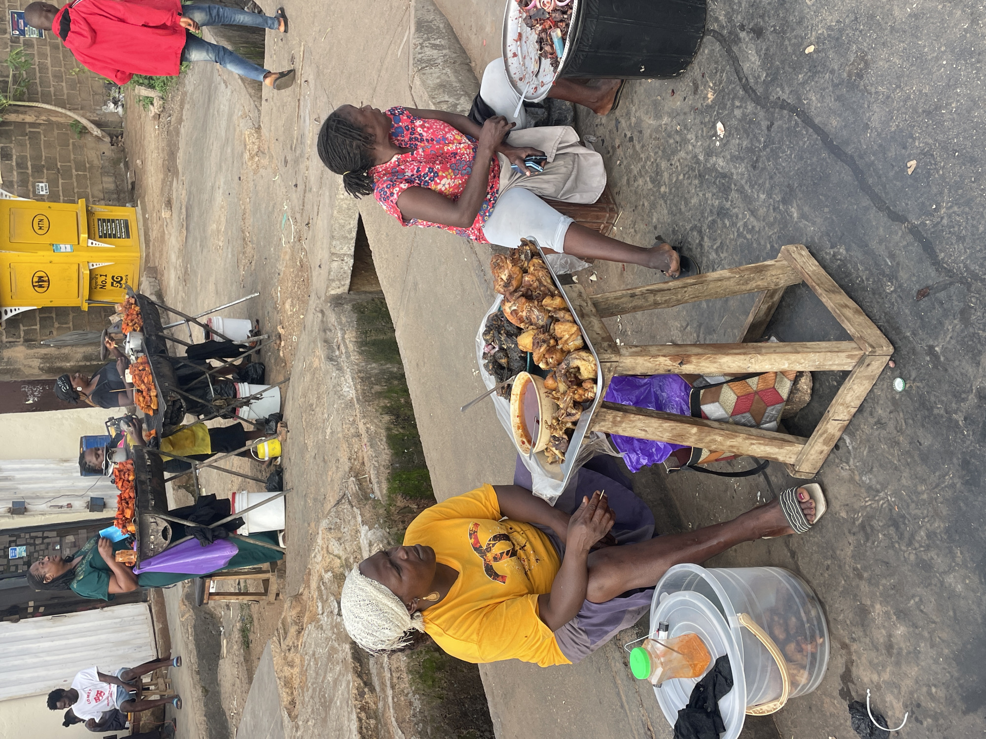 The image depicts an outdoor street food vending scene in Jos, Nigeria. In the immediate foreground, two women are seated on the paved ground. On the left, a woman with dark skin wears a white headwrap, a yellow short-sleeved t-shirt with a black and red "CC" logo, and a dark purple skirt, seated with her legs bent. She wears open-toed sandals. To her right, a low wooden table holds a metal tray laden with cooked meat pieces (likely chicken) and a small bowl of dark red sauce. A purple plastic bag hangs from the table. To her left, a clear plastic bucket with a lid contains food, and a small plastic bottle with orange contents is connected by wires to a small black device. On the right, another woman with dark skin, wearing a red and blue floral sleeveless top and light-colored capri pants, sits on a low stool or crate. She holds a dark smartphone in her lap and wears black flip-flops. A metal tray with cooked meat is to her left, and to her right, a black barrel-like container with a metal tray on top holds chopped meat with red onions.
In the mid-ground, a woman in a dark green dress stands behind a charcoal grill filled with skewered, cooked meat. She holds a light blue plastic bag. Adjacent to her, another charcoal grill with similar skewered meat is attended by a partially visible individual in a yellow shirt. Behind these grills, several individuals are seated outdoors, including a man in a white t-shirt and blue shorts on the far left.
The background features a building with light-colored roll-down shutters on the left and a brick wall on the right. Prominently displayed against the brick wall are two yellow MTN kiosks, marked "Nigeria's No. 1 5G Network." A man in a red hooded jacket and blue jeans walks away from the viewer towards the right of the frame. The ground throughout the scene is uneven, grey concrete with cracks and dirt patches, indicating a well-worn urban setting.