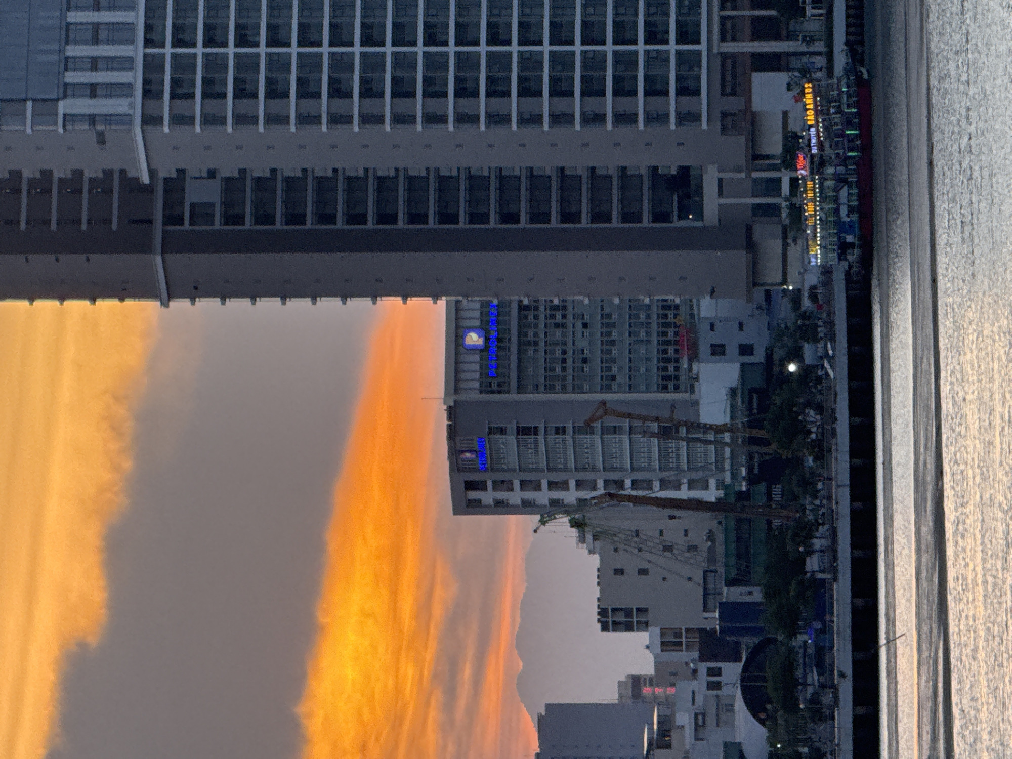 An aerial photograph captured at dusk shows a wide, calm body of water in the foreground, reflecting the warm orange and yellow hues of the sky. Beyond the water, a city skyline rises, dominated by modern high-rise buildings. To the left, a smaller, lighter-colored building stands with several smaller buildings clustered around its base. In the center, a taller, gray building displays a blue logo that reads "PETROLIMEX." To its right, an even taller, gray building with a grid-like pattern of windows dominates the frame. Cranes and other construction equipment are visible between the buildings, suggesting ongoing development. In the distance, hazy mountains are visible against the sky. Along the waterfront, a brightly lit boat with multiple decks is docked, adorned with colorful lights and signage that appears to be in Vietnamese. Trees line the embankment in front of the buildings. The overall scene depicts an urban coastal environment at sunset.
